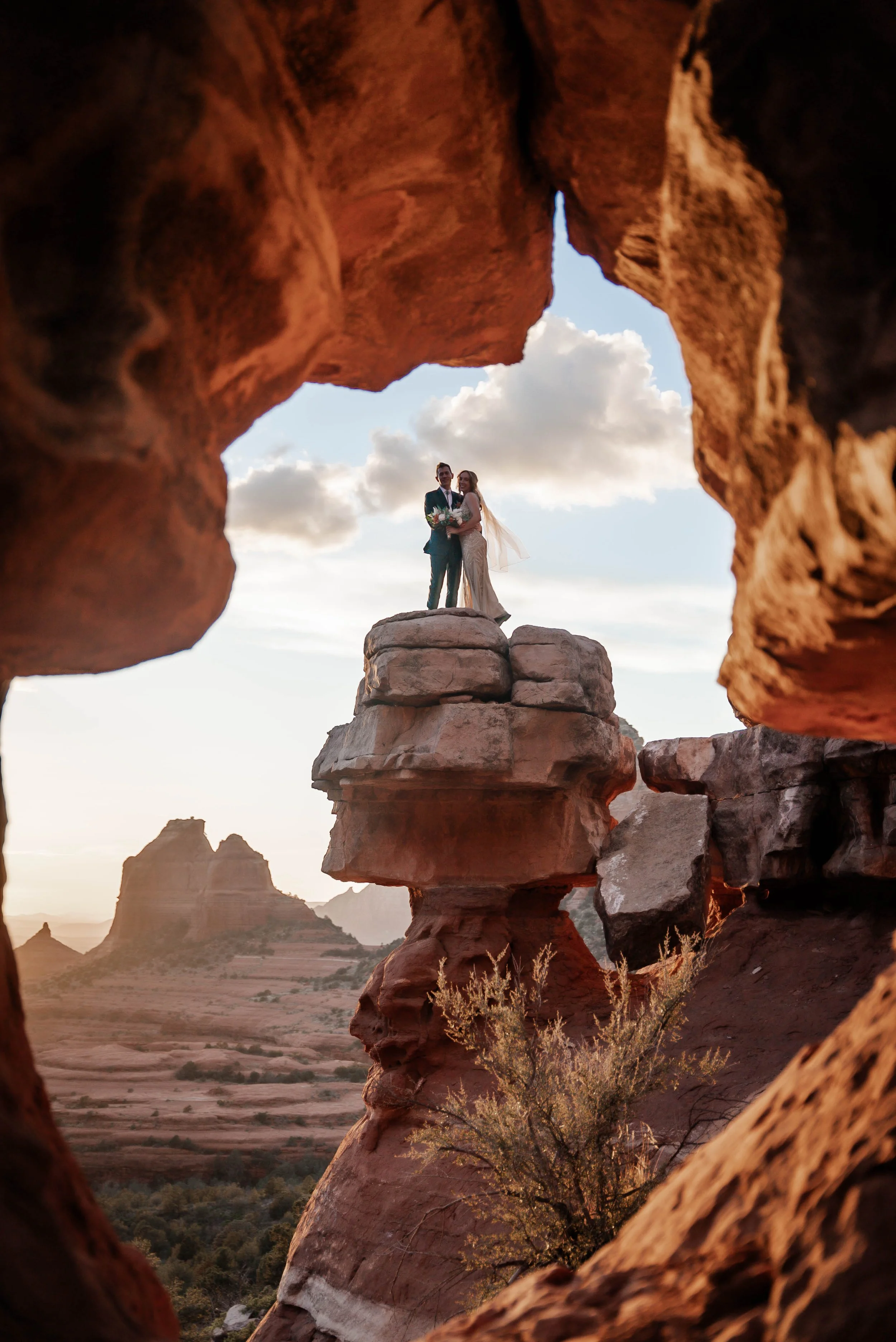 A bride and groom standing on a rock formation in a desert landscape, framed by rocks, during sunset with clouds in the sky.