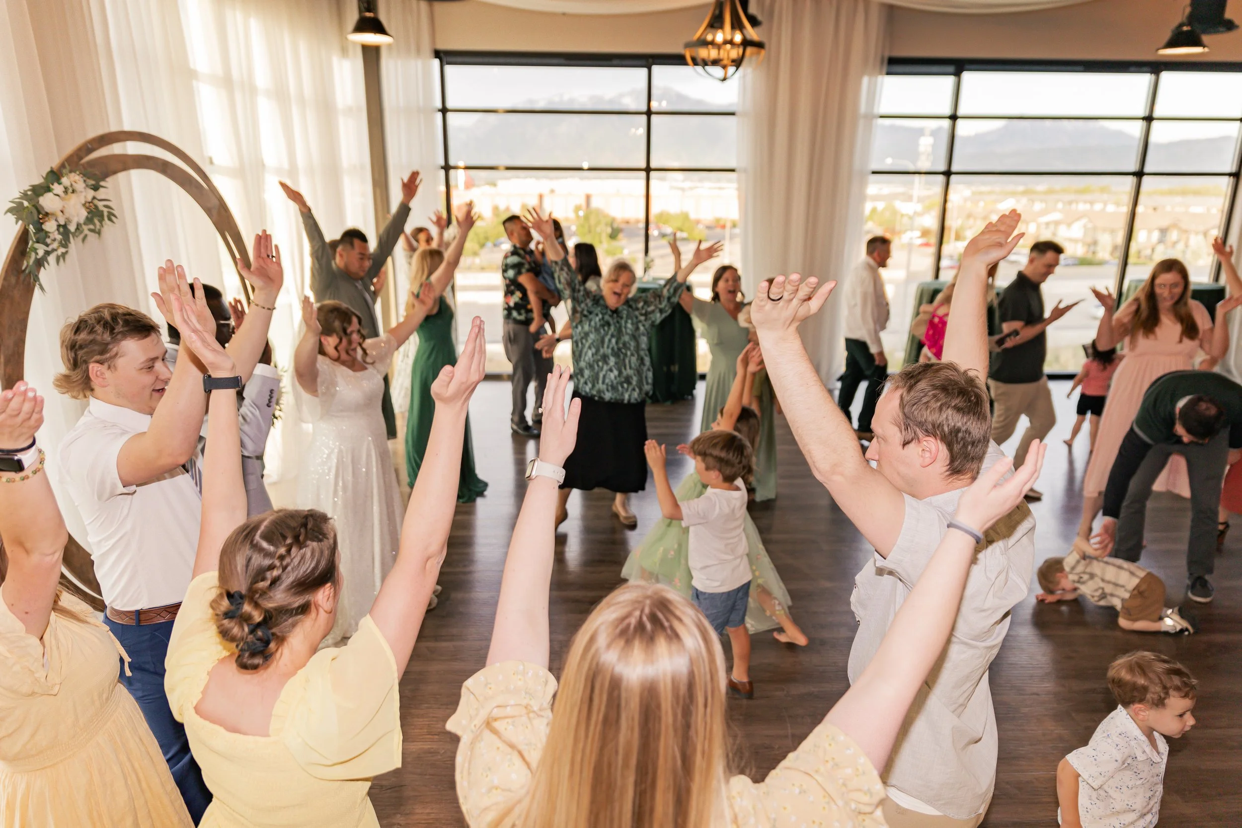 People dancing and celebrating at a wedding reception in a large, well-lit room with large windows and curtains, with children and adults enjoying the moment.