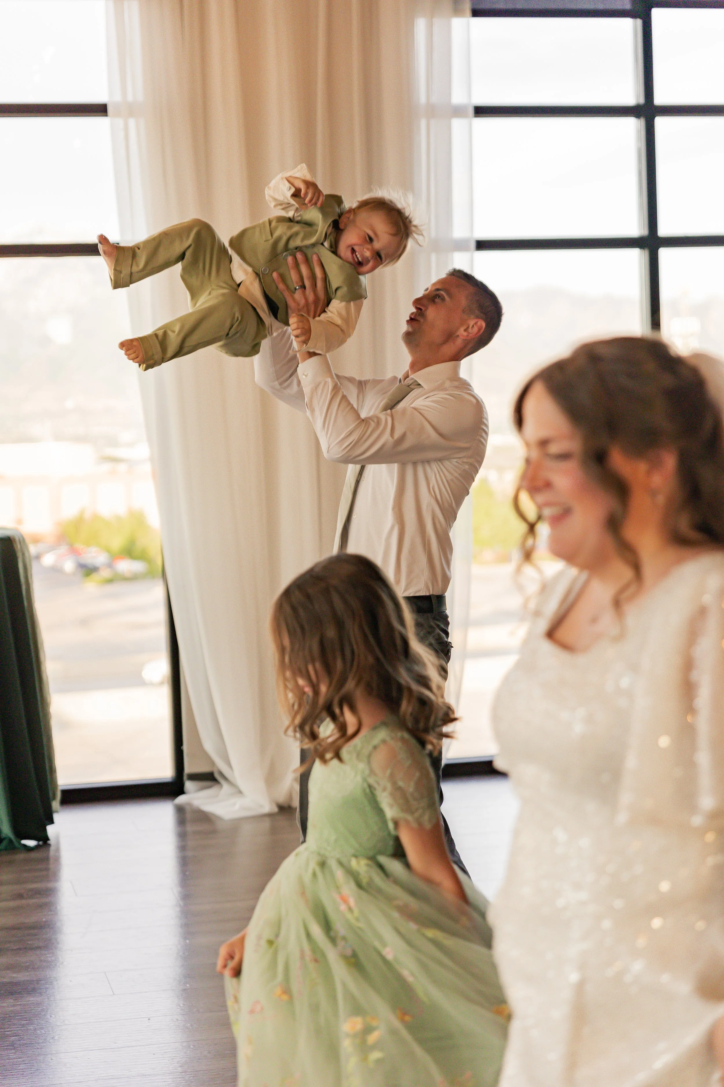 A man lifting a young girl in a light-colored dress, her arms raised and smiling, inside a room with large windows and curtains, with a woman and a young girl in the foreground.