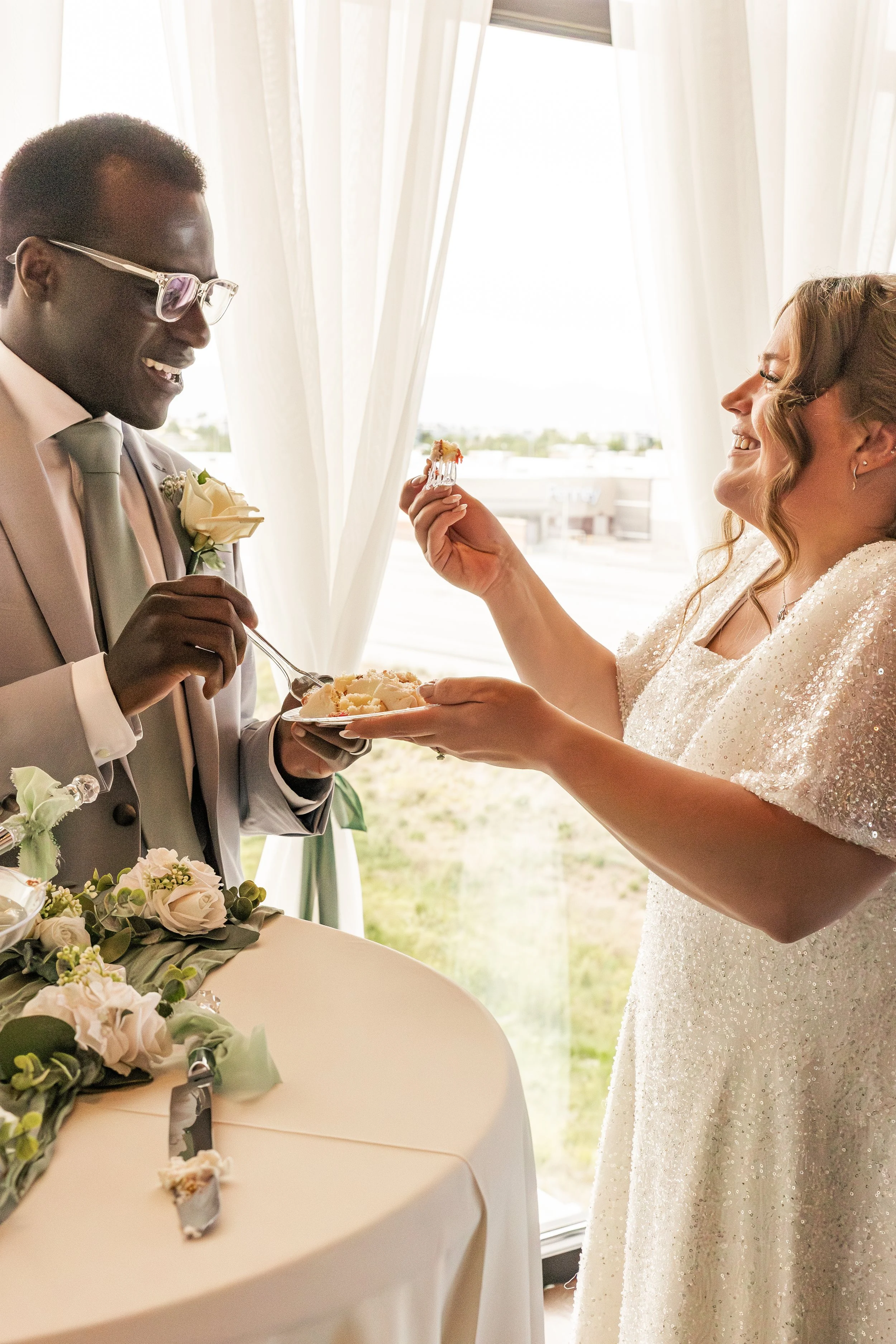 A bride feeds cake to a groom during their wedding ceremony, with both smiling joyful. The groom is wearing a light suit and glasses, and the bride is in a white, glittery dress. There is a table with flowers and a cake knife in front of them, and na