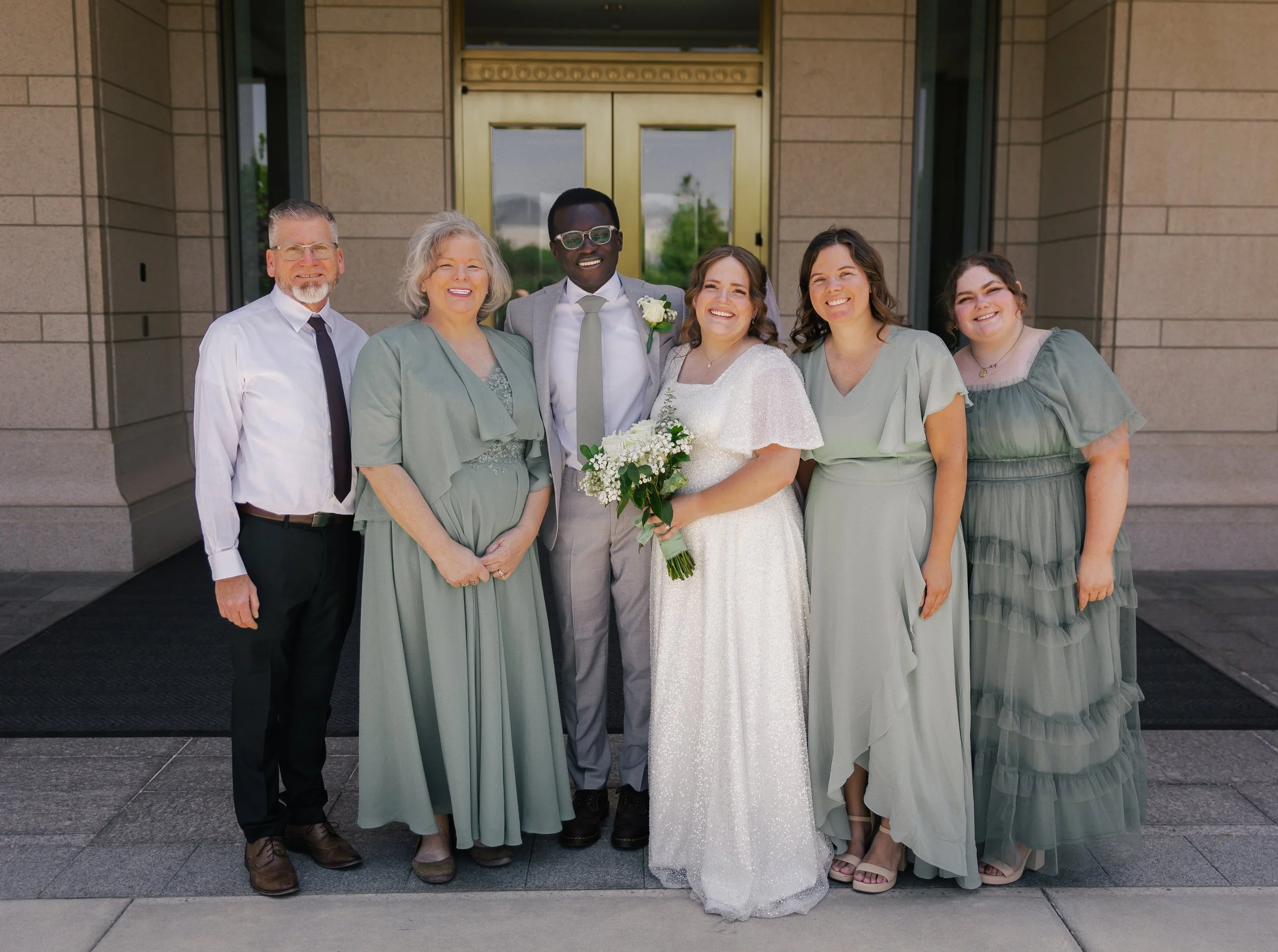 Group of six people standing in front of a building, dressed in wedding attire, smiling at the camera. The bride is holding a bouquet of white flowers, wearing a white gown. The groom is wearing a light gray suit with a white shirt and tie. Accompany