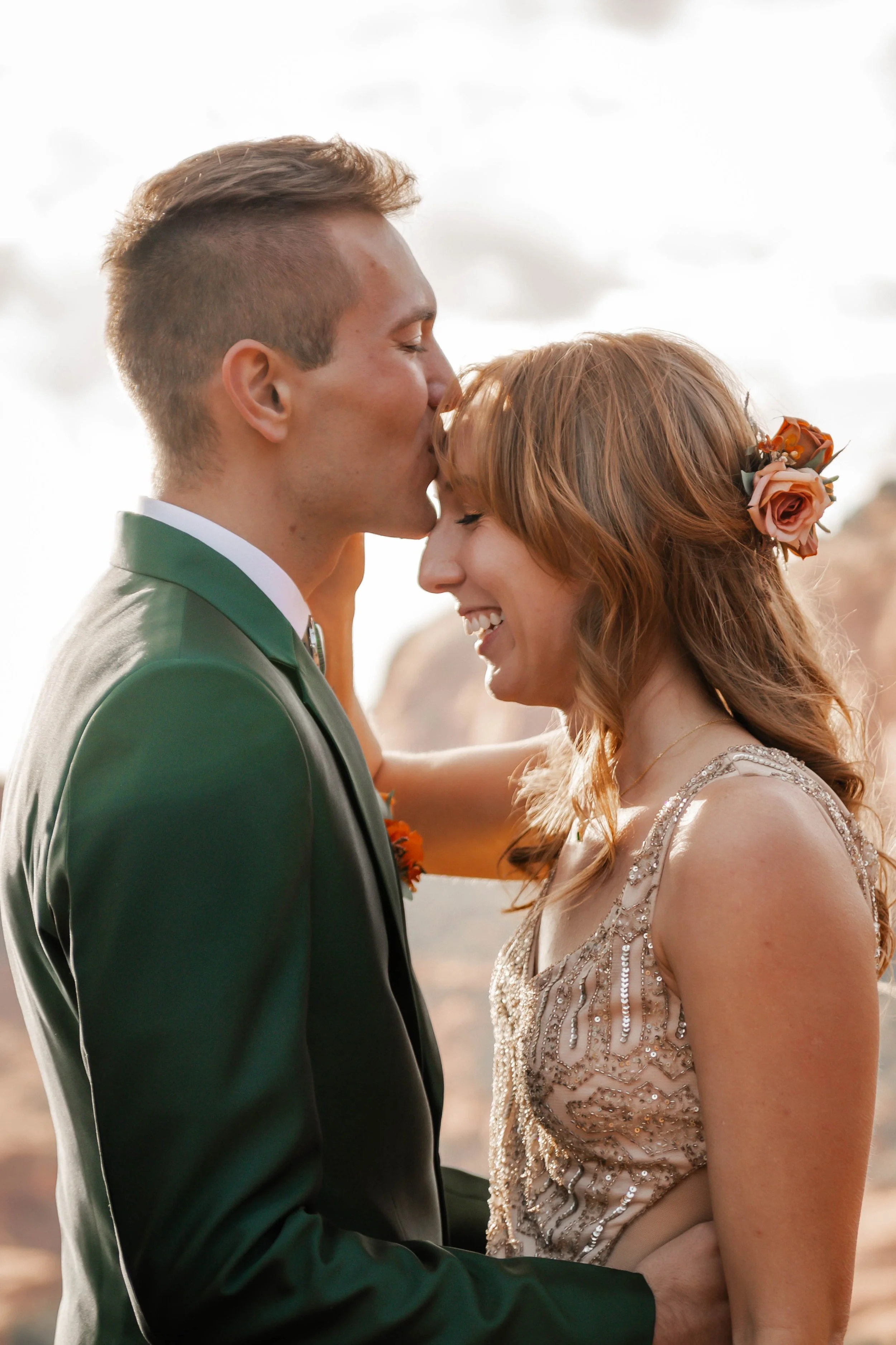 Couple at their wedding, with the groom kissing the bride on the forehead, outdoors with a bright sky and rocky background.