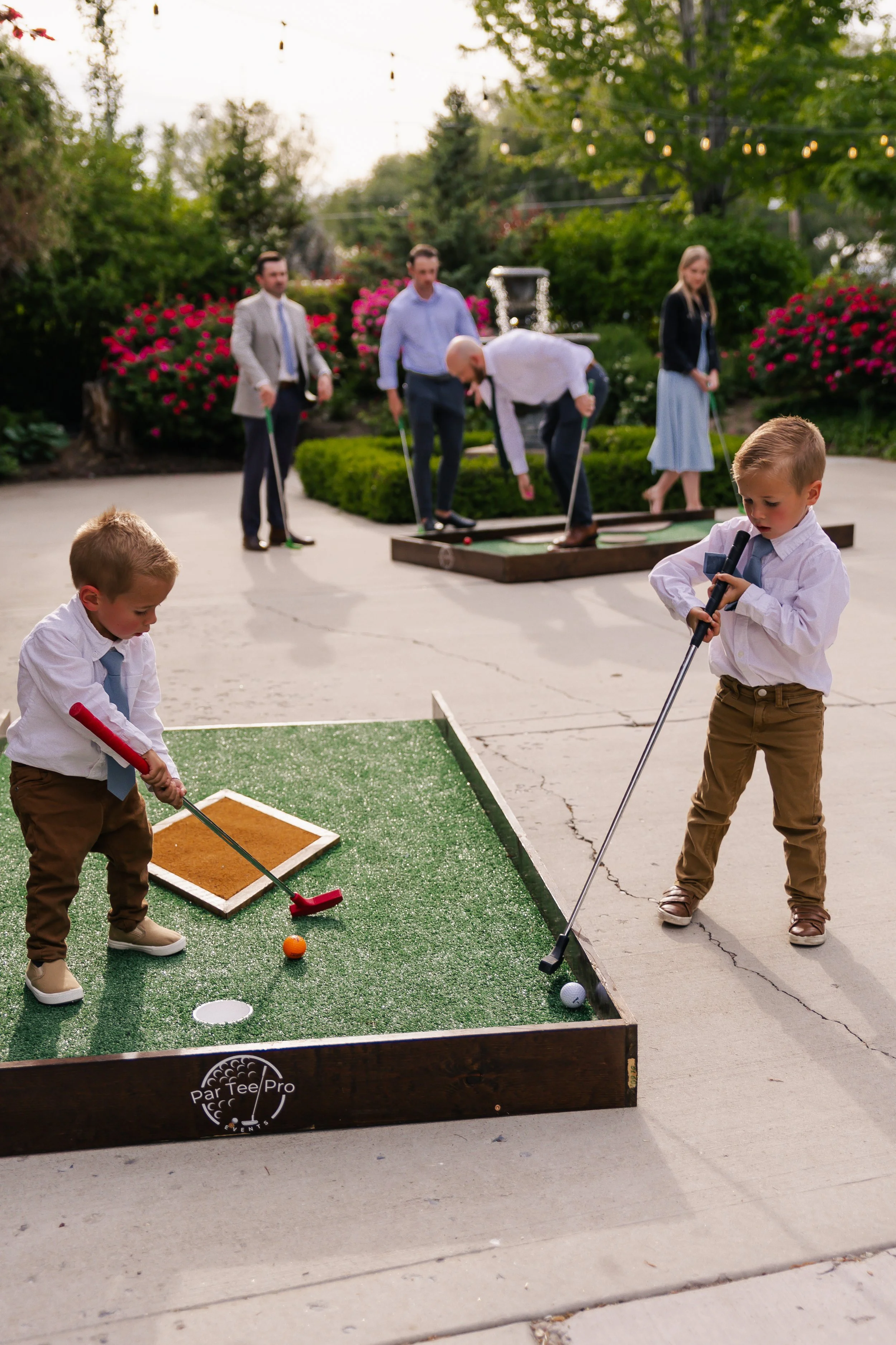 Two young boys playing miniature golf on a green turf course with adults playing in the background near a garden with pink and purple flowers; they are dressed in formal attire.