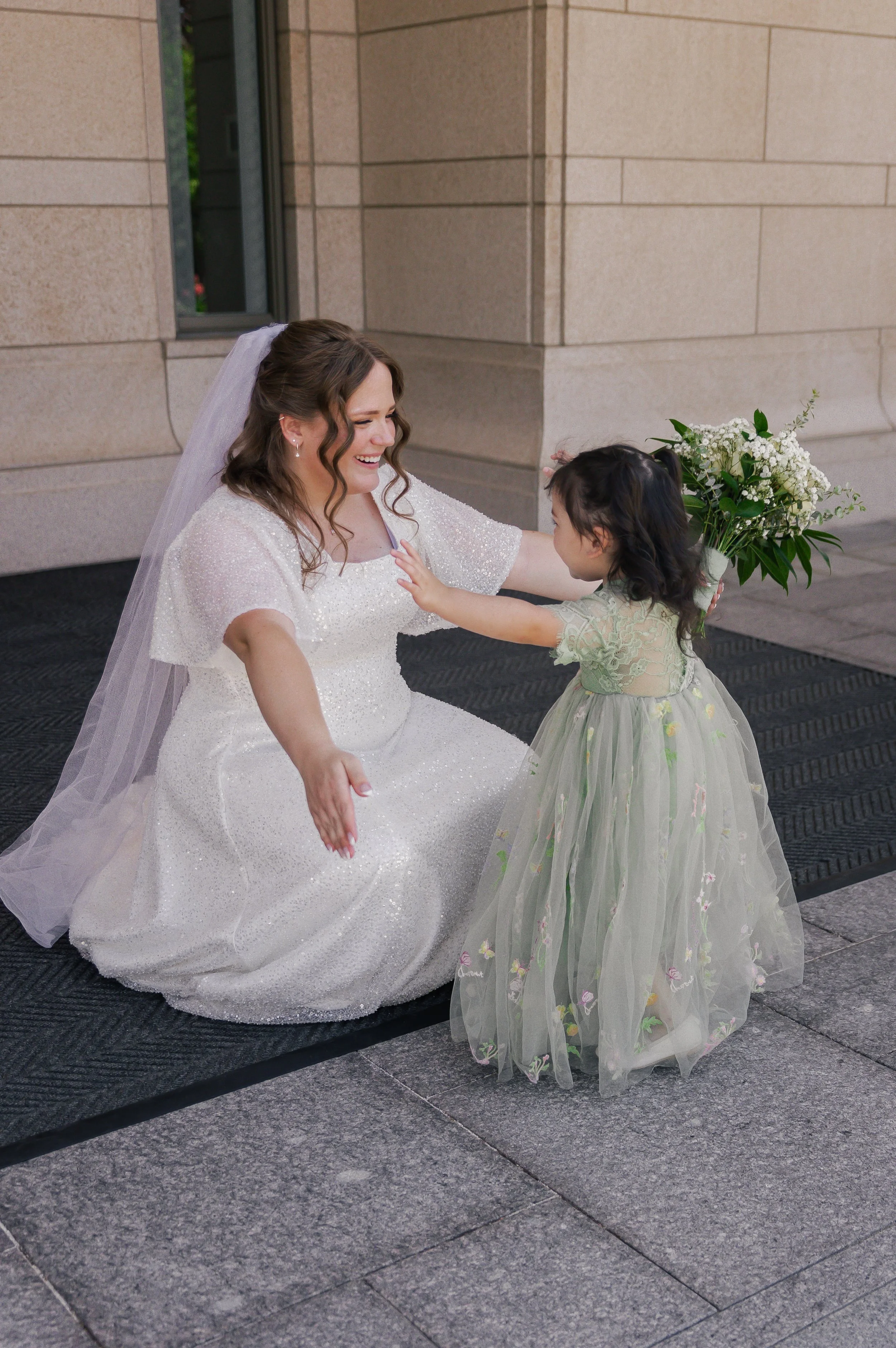 A bride in a white wedding gown smiling and squatting down, reaching out to a young girl in a pastel green dress, who is holding a bouquet of flowers outside of a building.