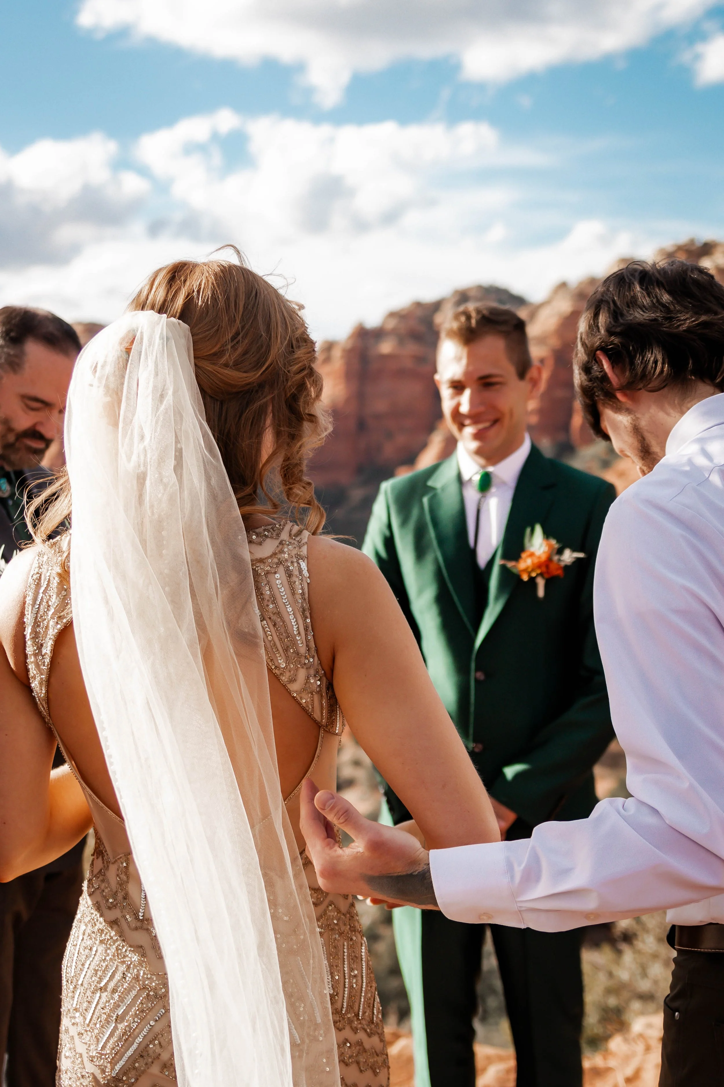 A wedding ceremony outdoors with a bride in a beaded gown and veil, a groom in a suit with a green lapel pin, and attendants, set against rocky red cliffs and a blue sky.