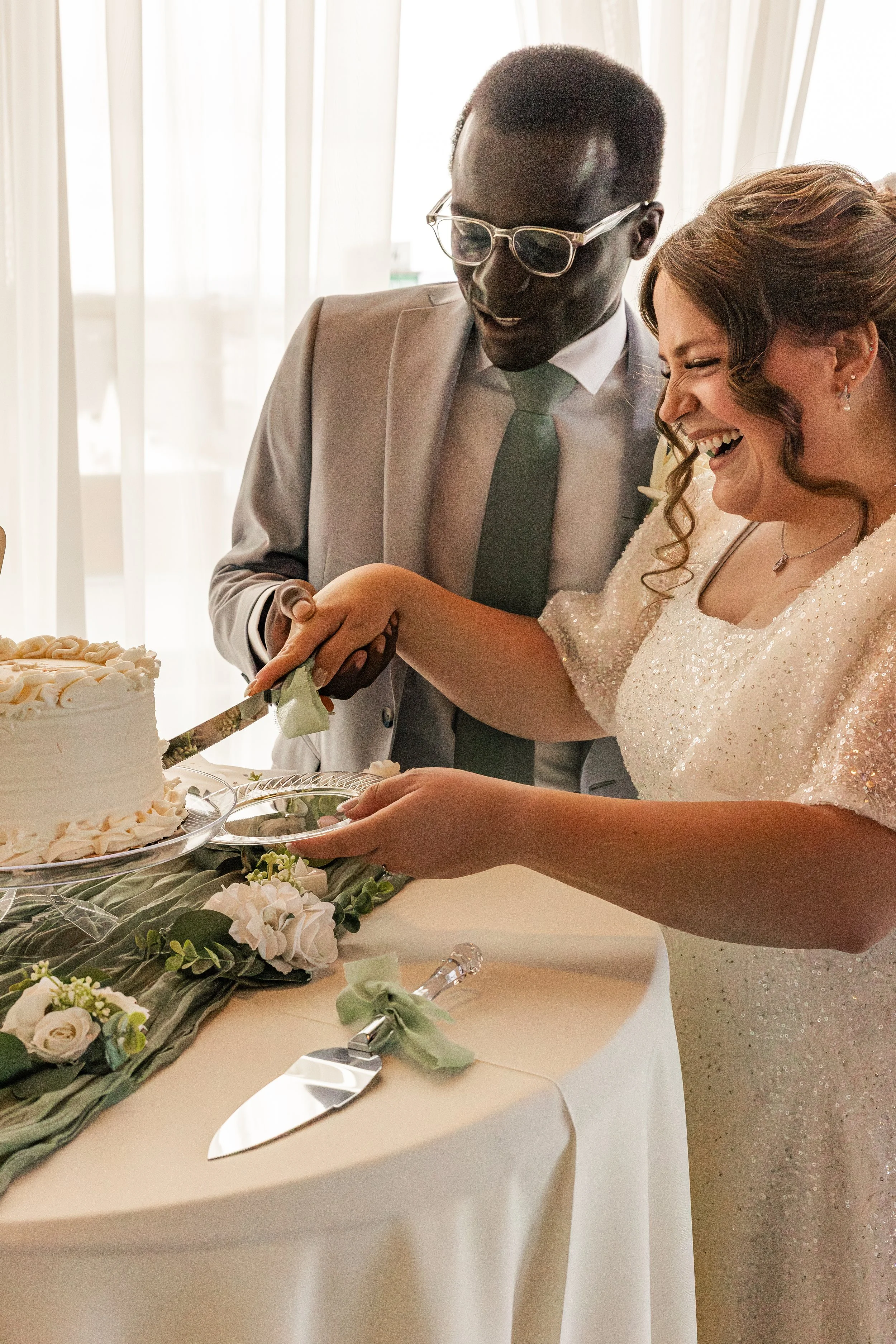 A bride and groom cutting their wedding cake together, smiling, with the bride wearing a white, sequined wedding dress and the groom in a gray suit and glasses.