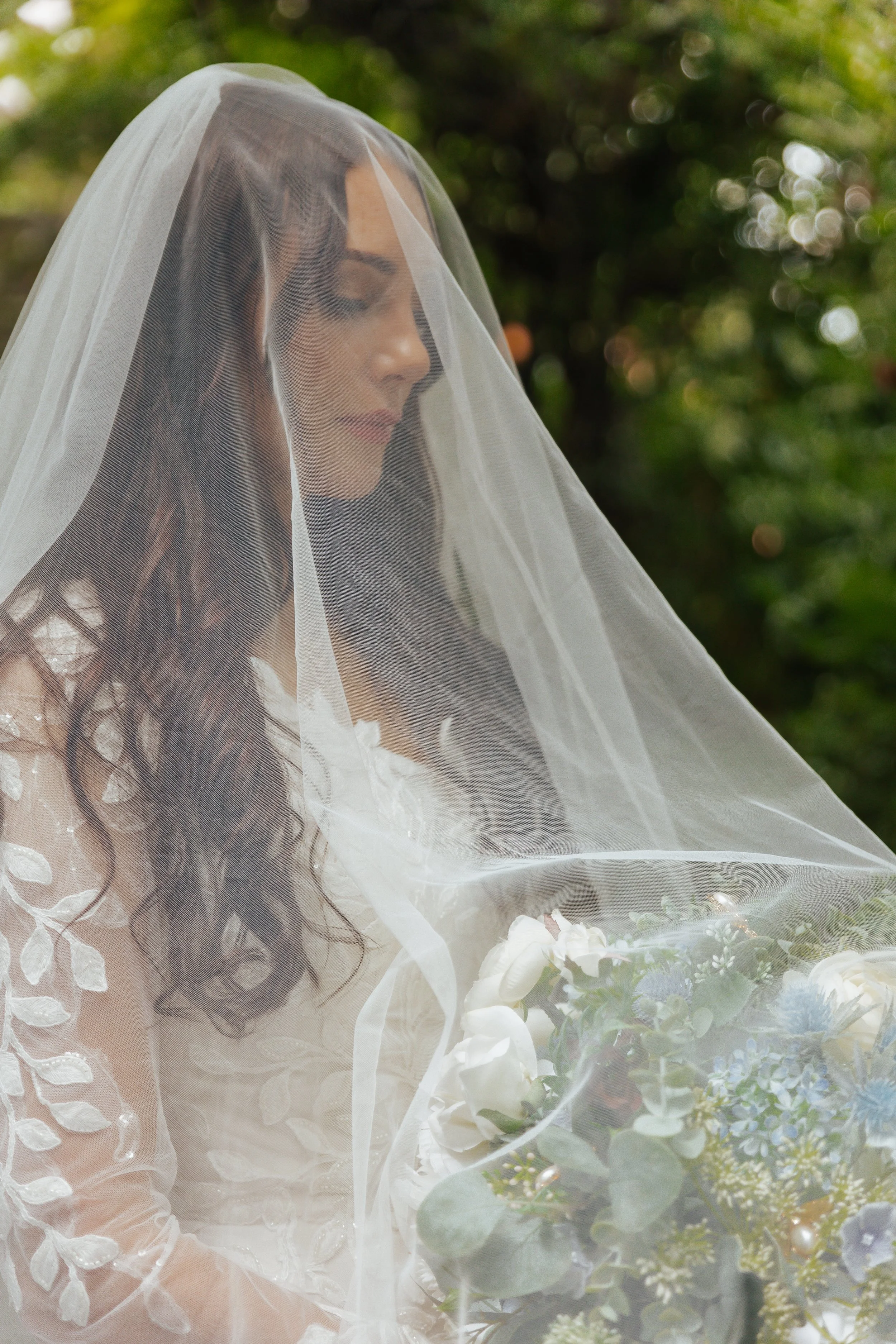 A bride with long, wavy brown hair wearing a white dress with floral embroidery, sitting outdoors with a bouquet of white, purple, and green flowers, and a sheer veil covering her face.