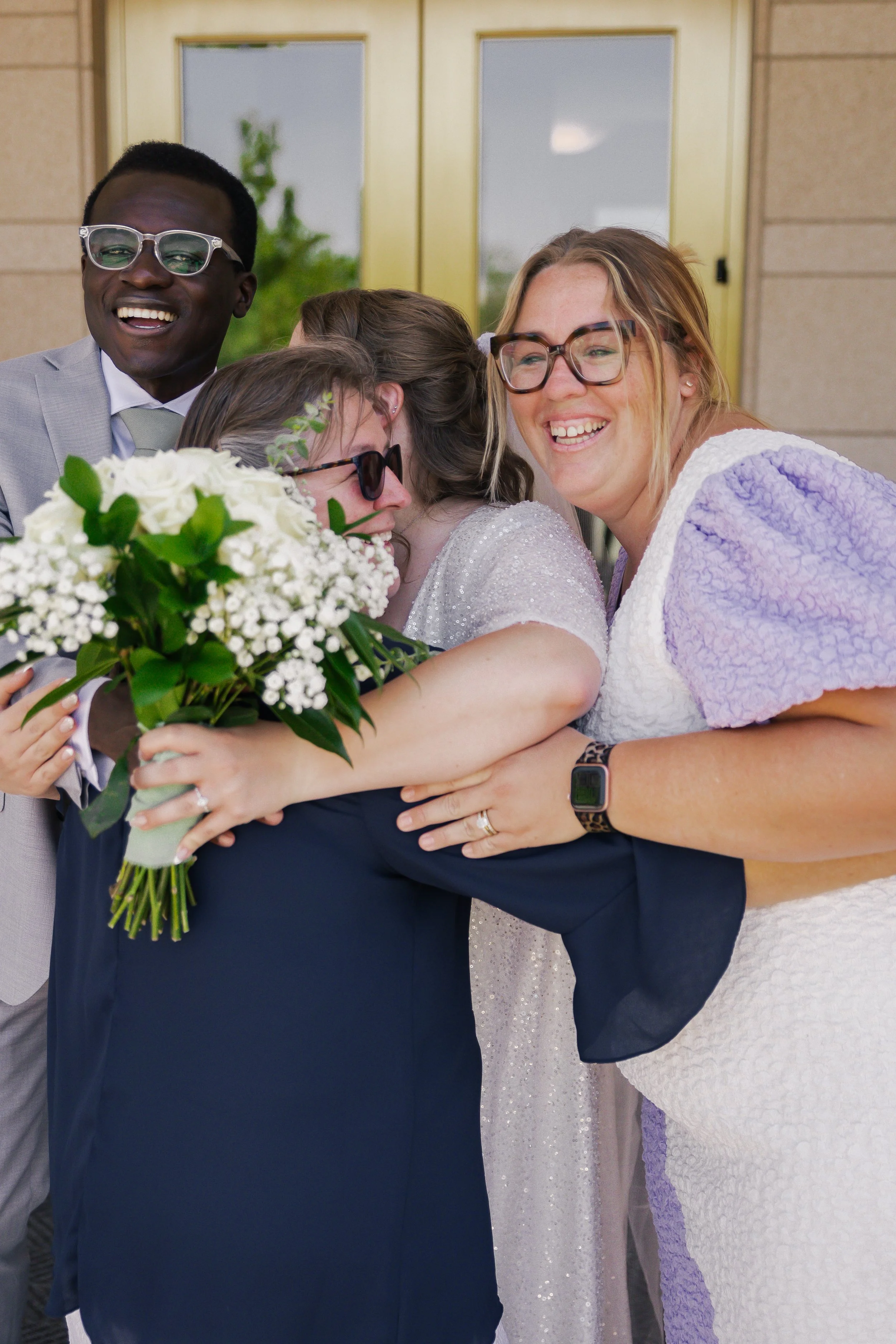 Group of joyful people hugging, with one holding a bouquet of white flowers, outside in front of a building entrance.