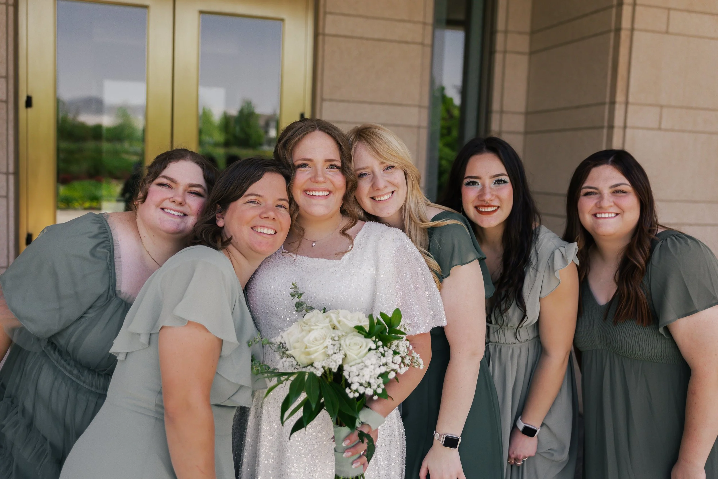 A bride in a white dress holding a bouquet of white roses, surrounded by five bridesmaids in green dresses, standing outside a building with glass and brick walls.