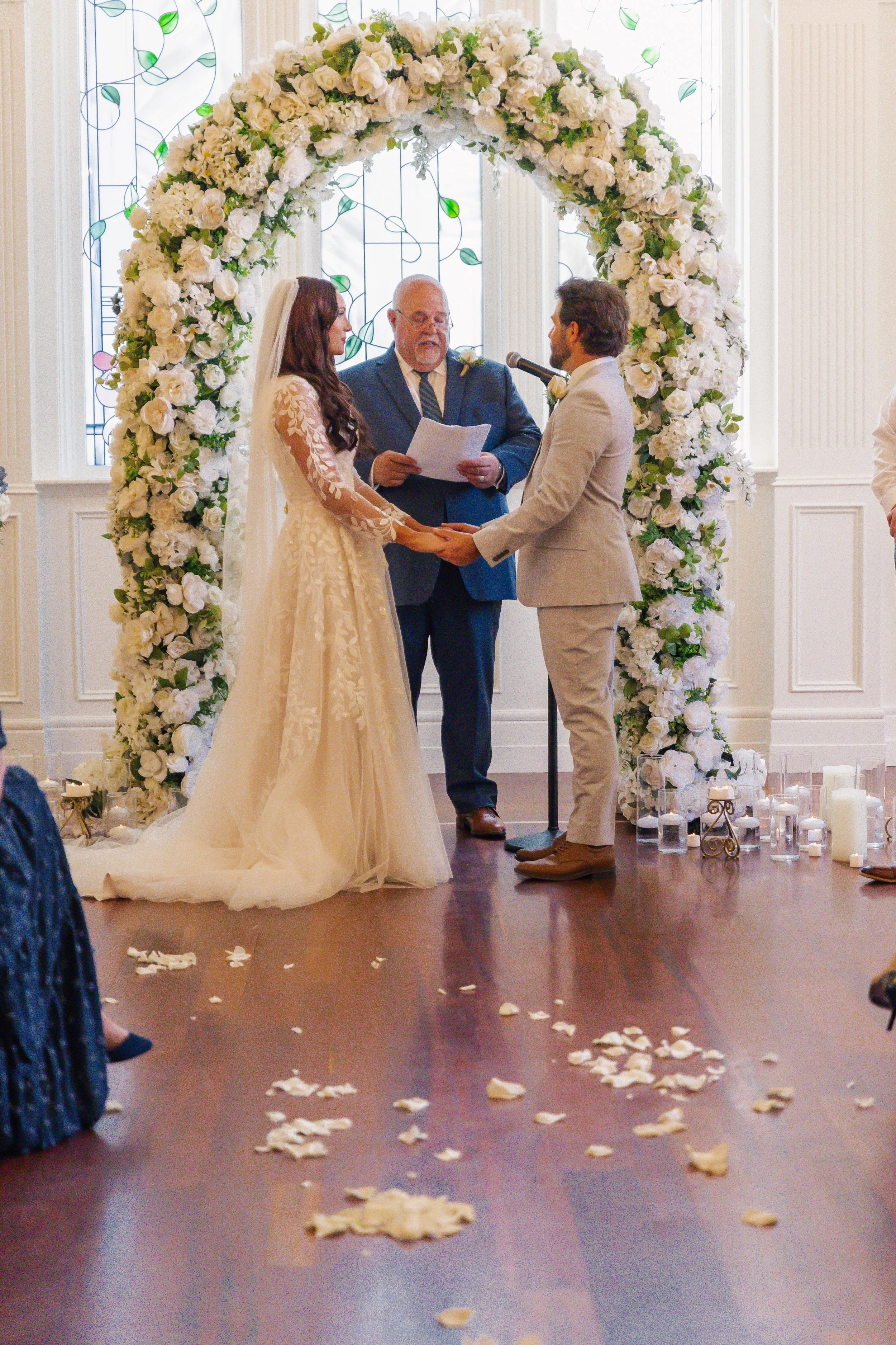 A wedding ceremony with a bride and groom holding hands under a floral arch, with an officiant reading vows, inside a decorated room with candles and rose petals on the floor.