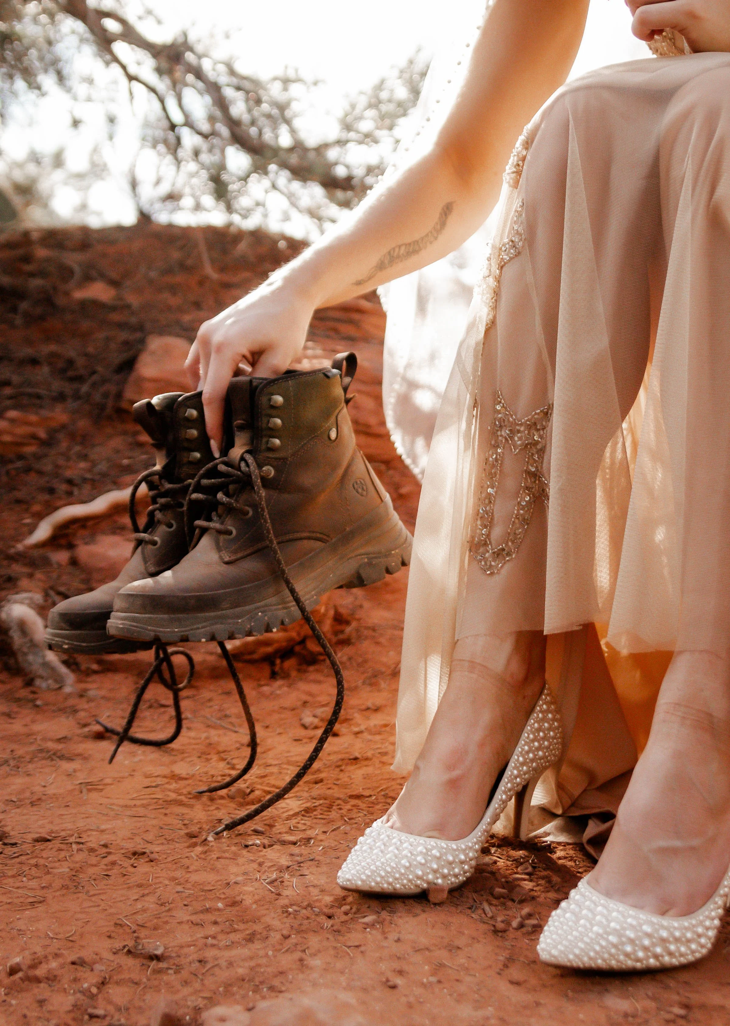A woman in a wedding dress holding a pair of brown combat boots outdoors on a red dirt ground with trees in the background.