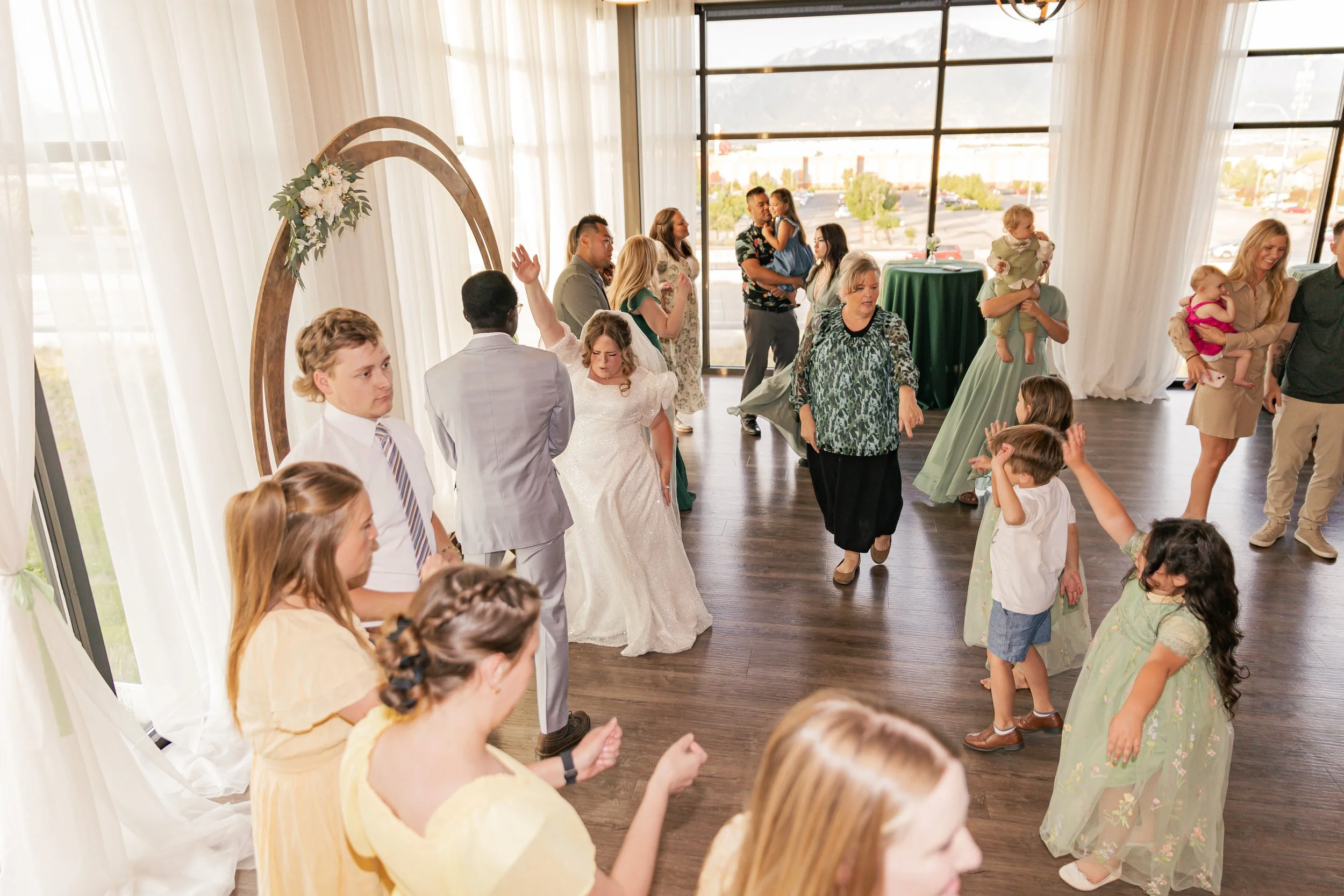People dancing and socializing at a wedding reception in a decorated indoor venue with large windows.
