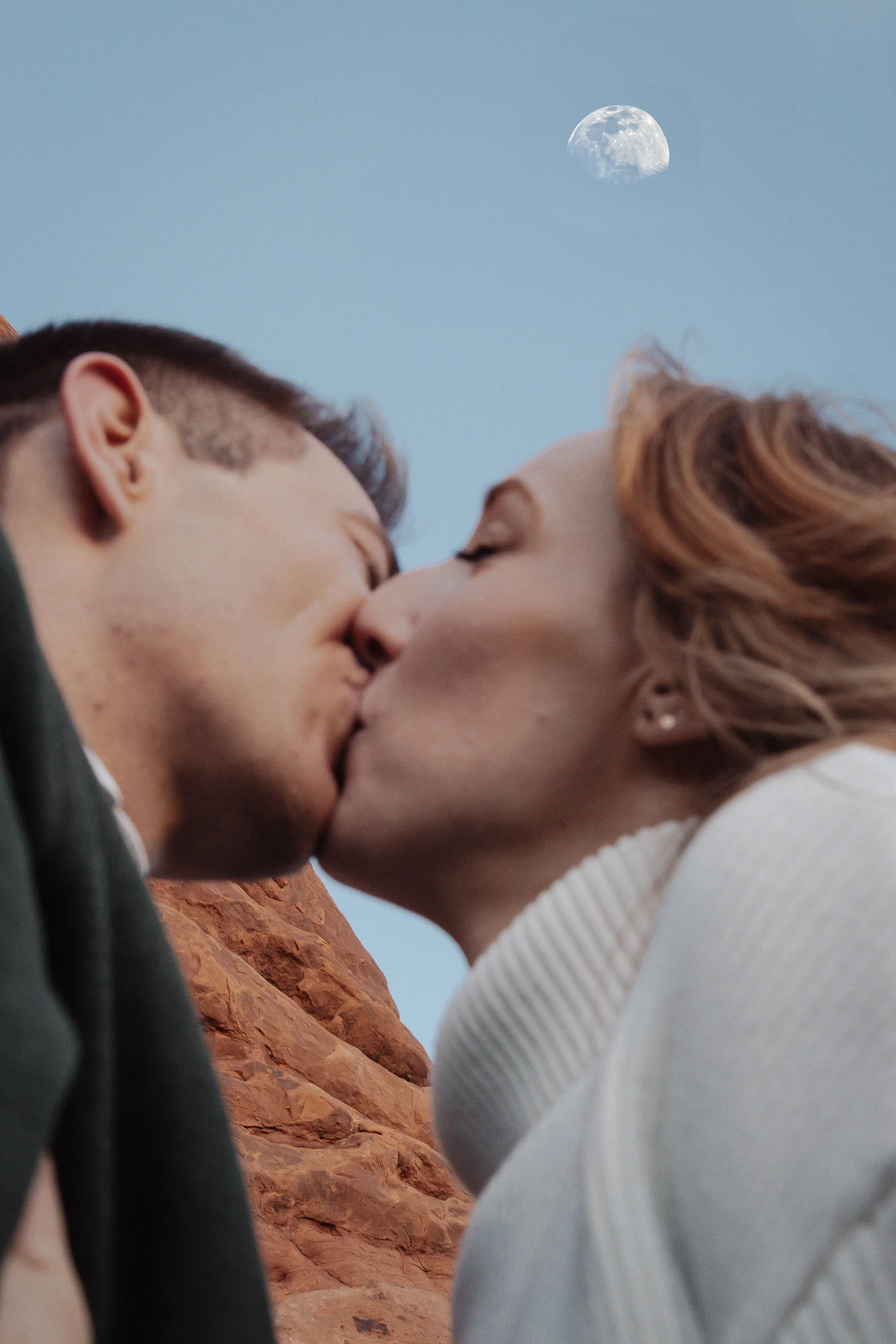 A man and woman are kissing outdoors, a red rock formation and the moon are visible in the sky above them.