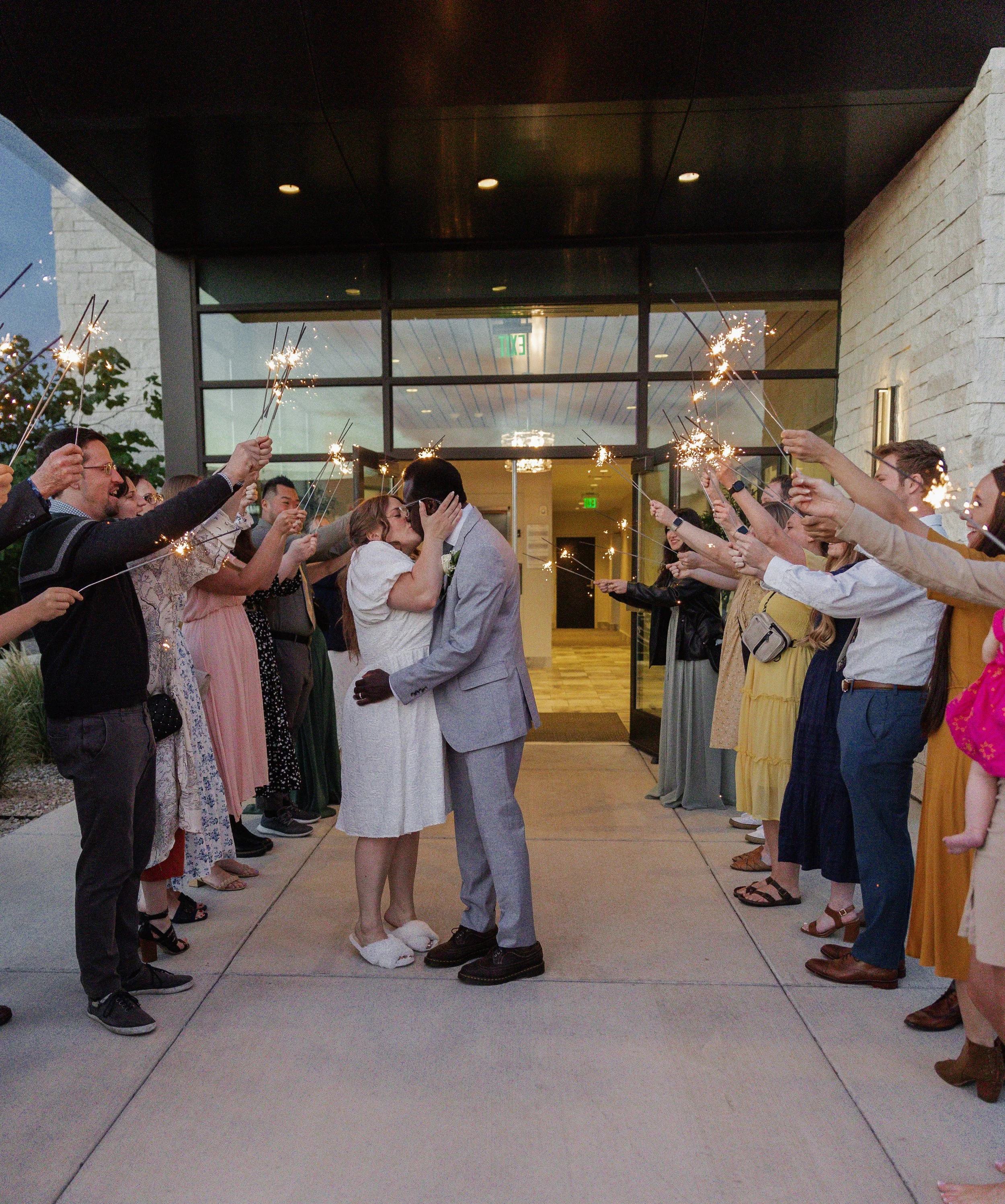 Couple kissing under sparklers held by friends at a wedding celebration outside a building.
