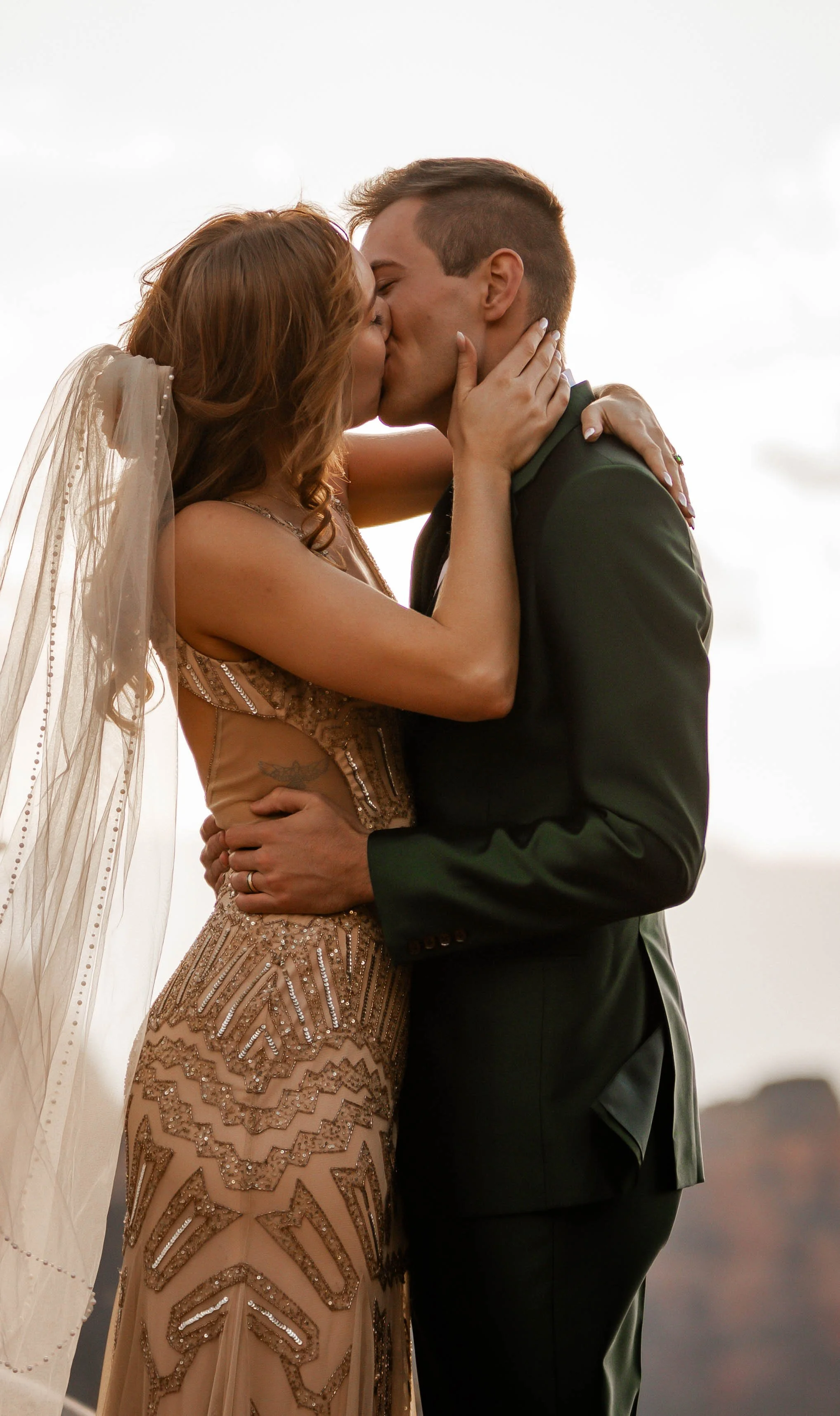 A bride and groom kissing outdoors, with the bride wearing a detailed, beaded gown and veil, and the groom in a dark suit.