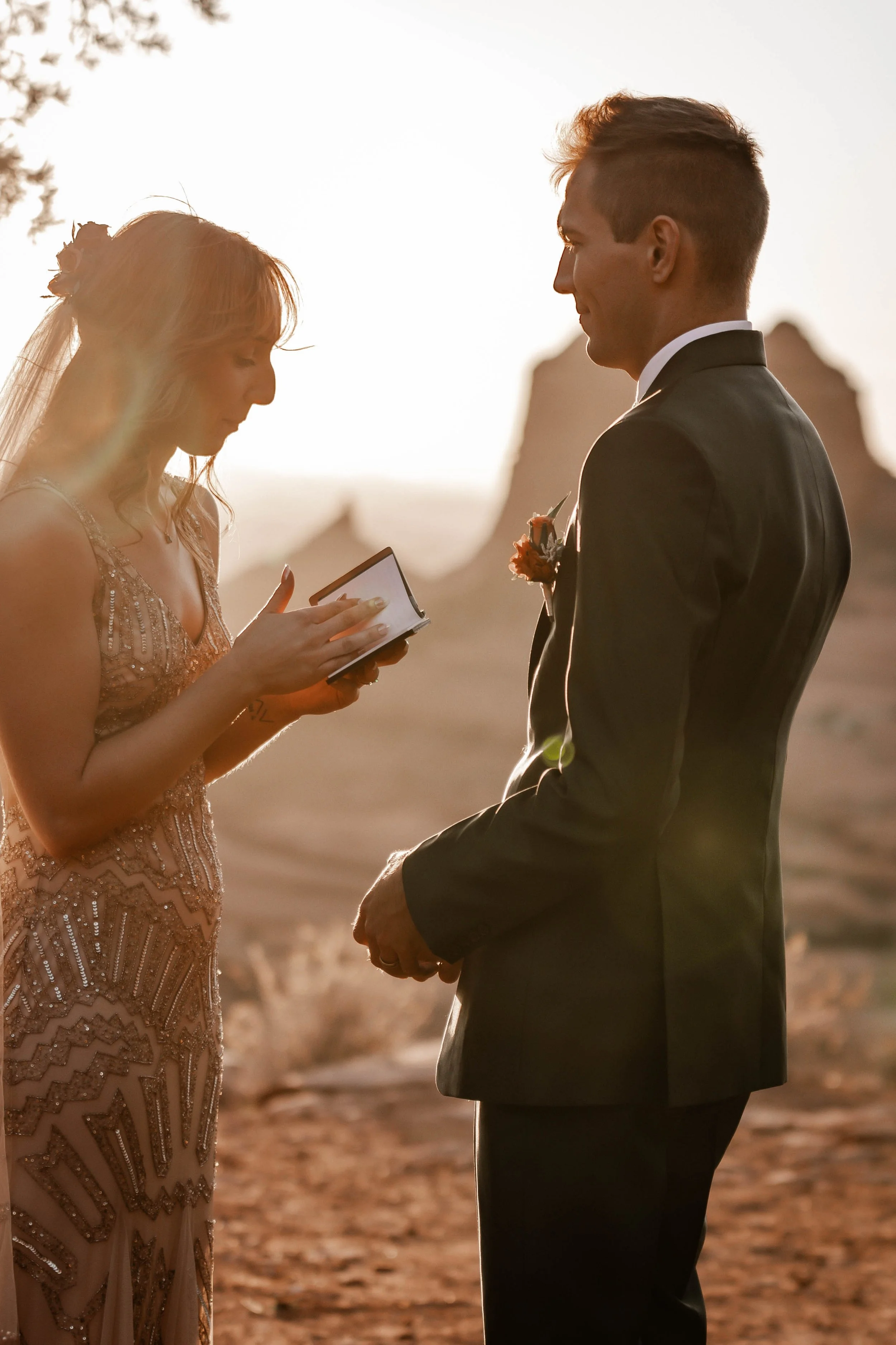 A bride and groom stand facing each other outdoors during sunset, with the bride holding a small book or paper, likely exchanging vows or rings.