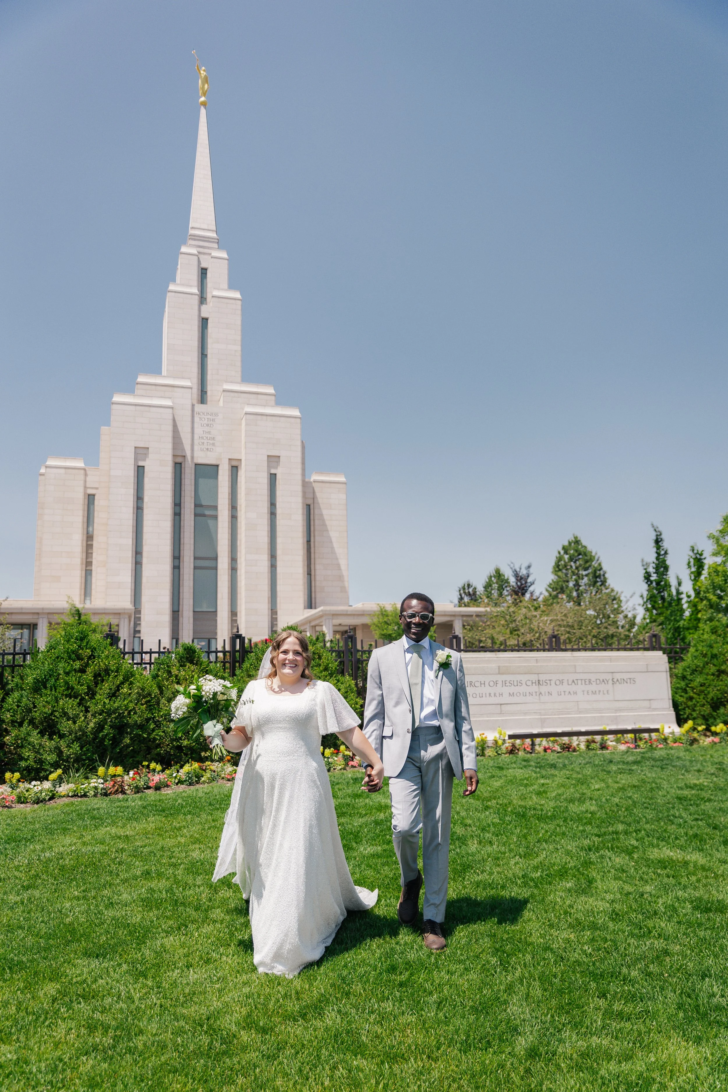 A bride and groom walking hand-in-hand outside a church, surrounded by green grass and flowers, on a sunny day.