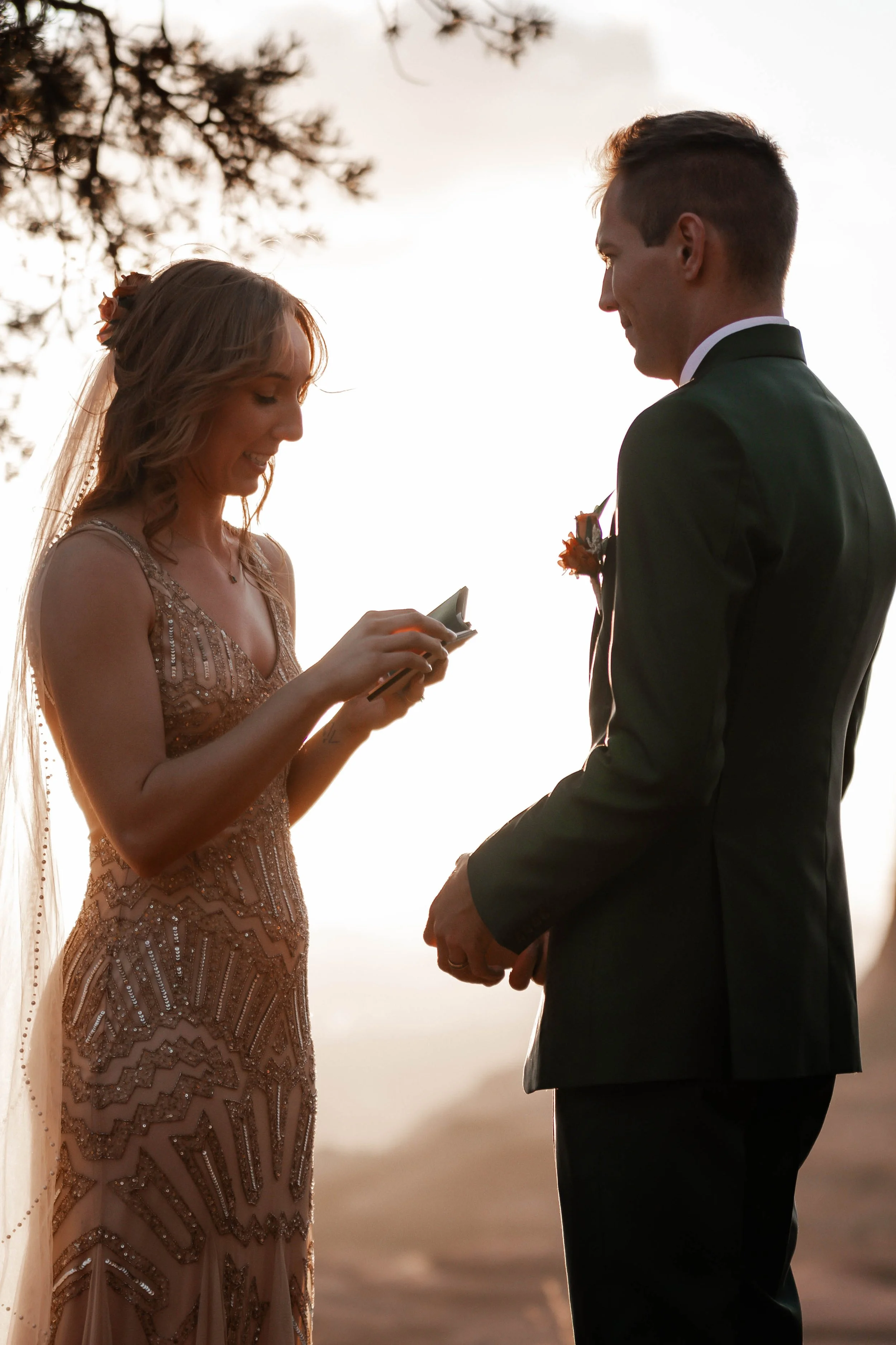A bride and groom exchanging vows during their wedding ceremony outdoors at sunset, with the bride reading her vows from a phone.