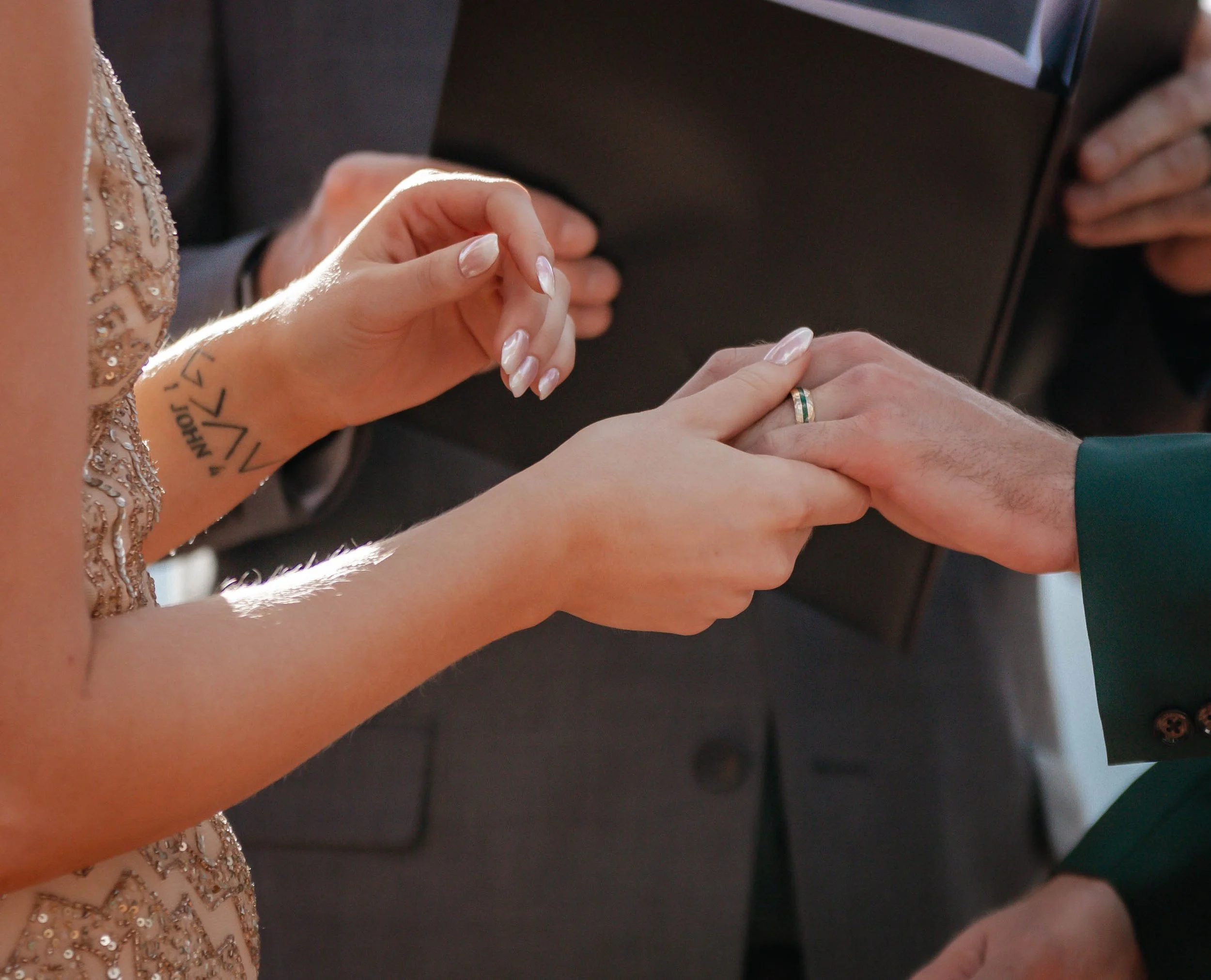 A woman wearing a sparkly dress holds a man's hand during a wedding ceremony, with an officiant holding a black folder in the background.