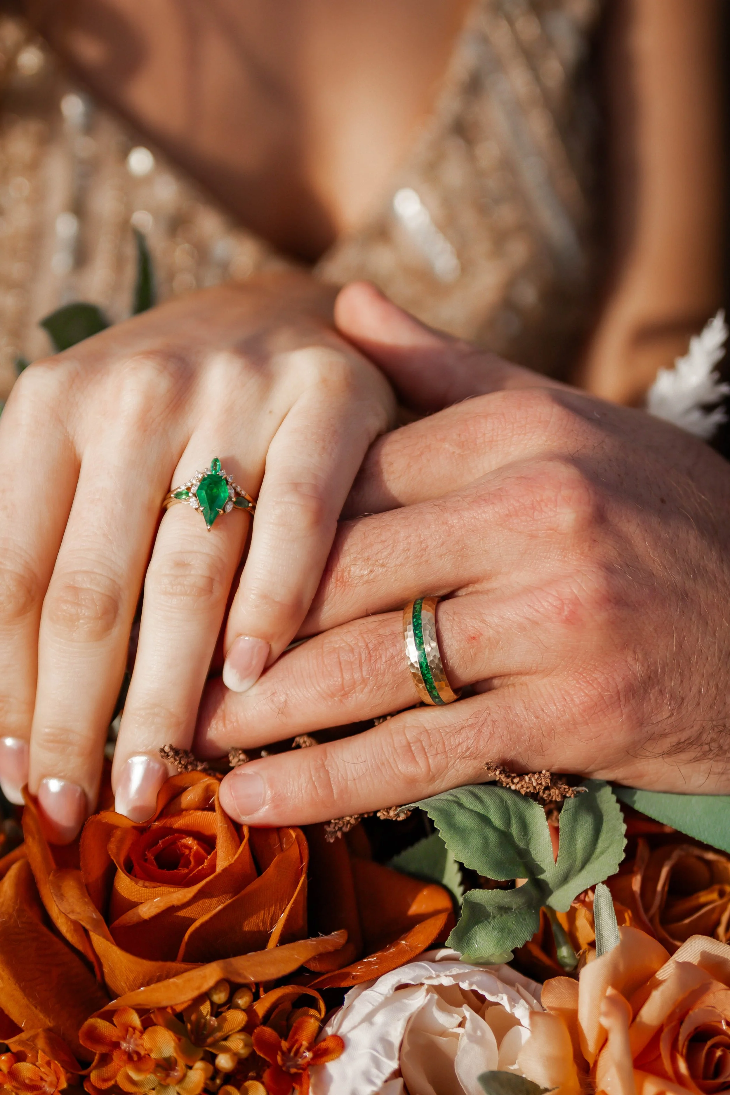 Close-up of a couple's hands with wedding rings, holding a bouquet of orange and cream flowers. The woman's hand features a green emerald and diamond ring, while the man's hand wears a silver ring with green accents.