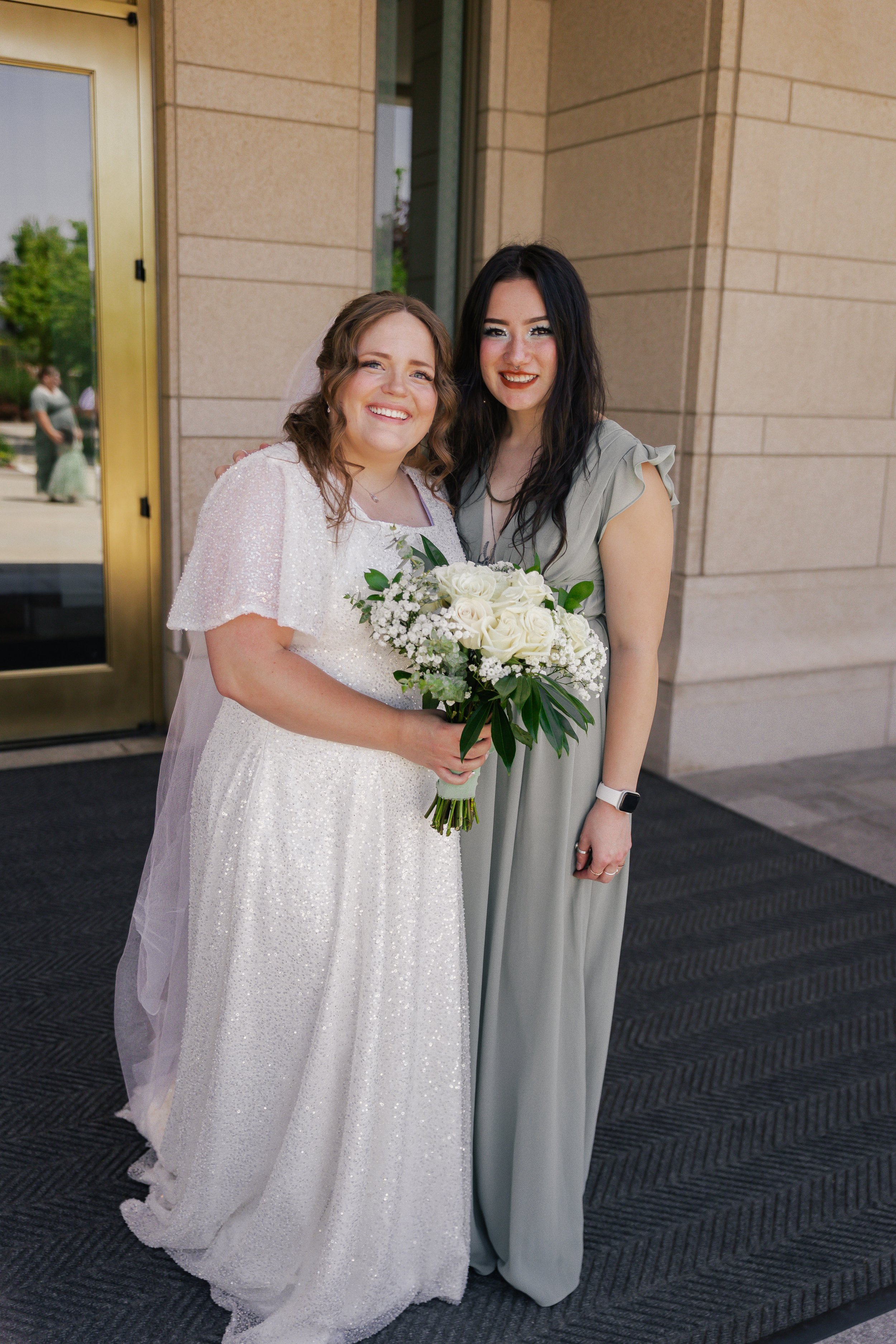 Two women, one in a wedding dress holding a bouquet, standing outside a building with a glass door and beige brick wall.