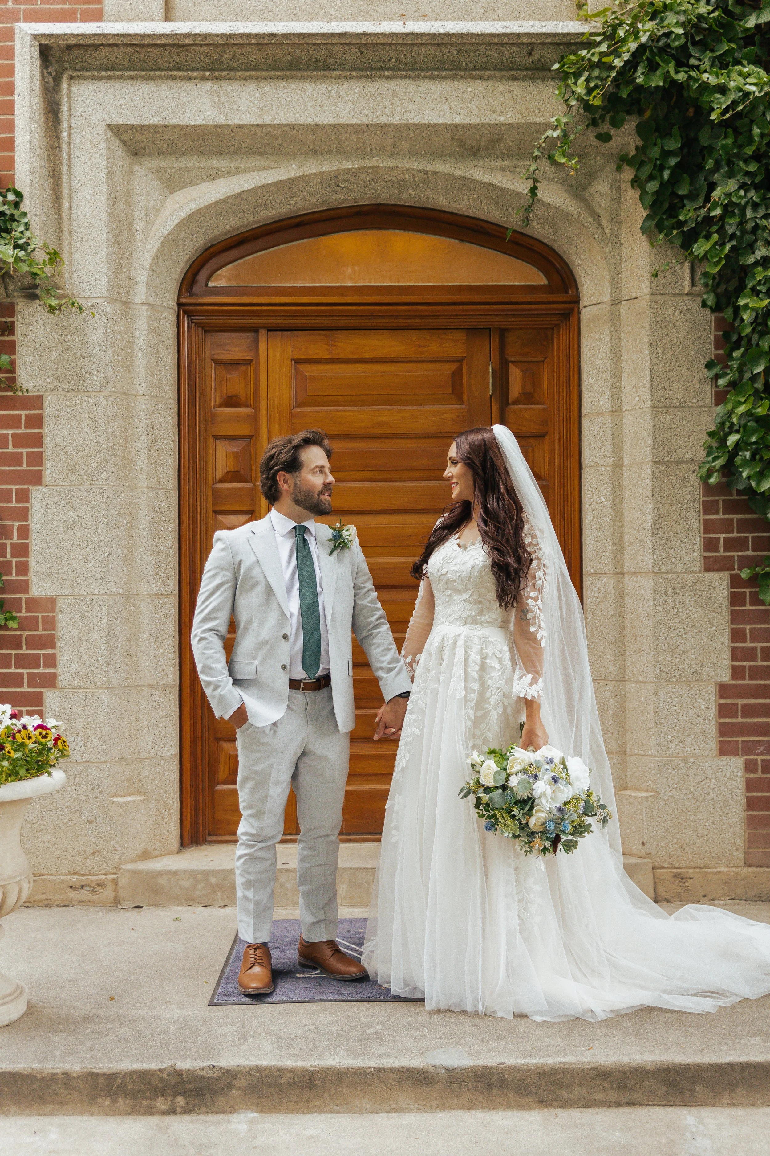A bride and groom holding hands outside a building with a wooden door, wedding attire, and a bouquet of flowers.