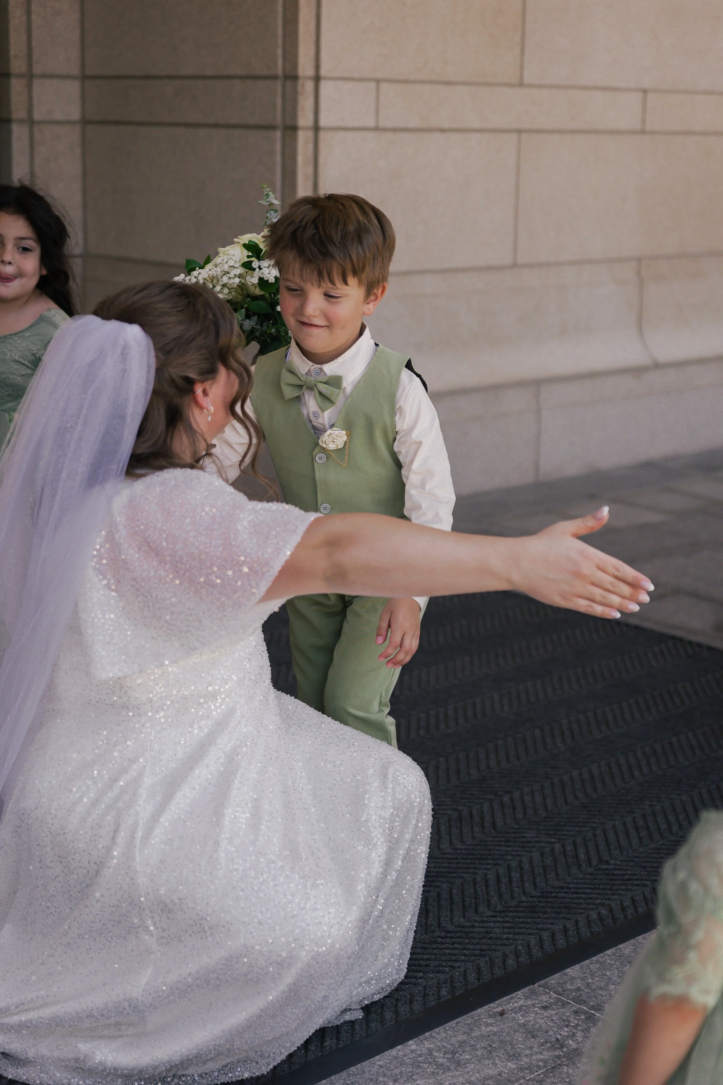 A bride dressed in a white gown is kneeling with her arm extended, giving a high five to a young boy in a green vest and bow tie at a wedding reception or ceremony.