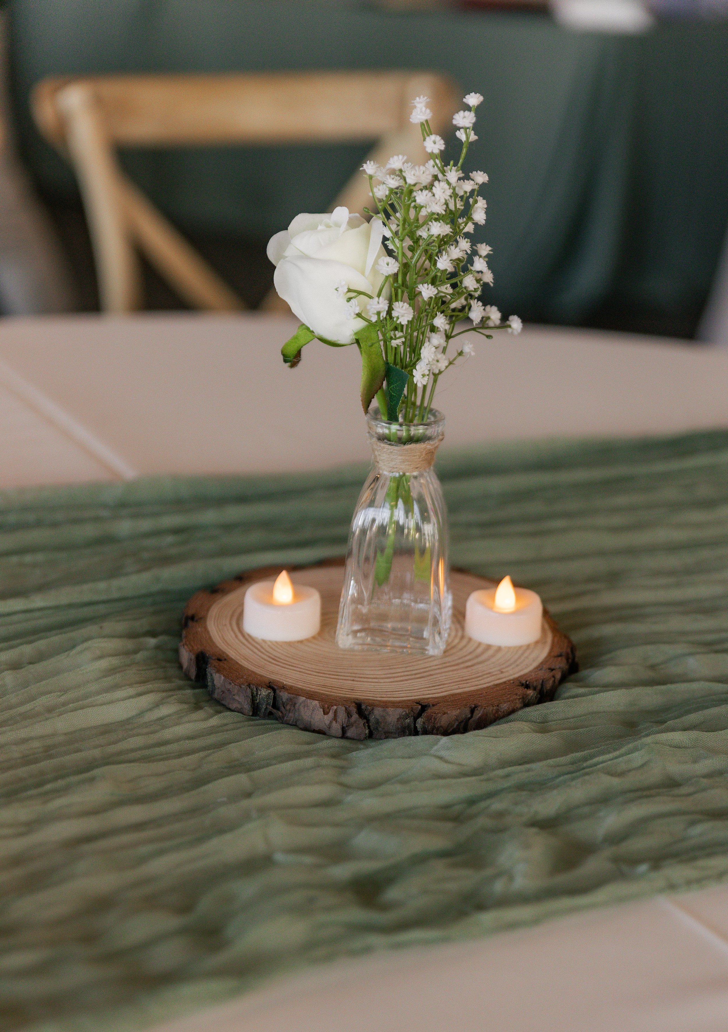A glass vase with white flowers and greenery, sitting on a wooden round slab, flanked by two small flickering candles on a green table runner.