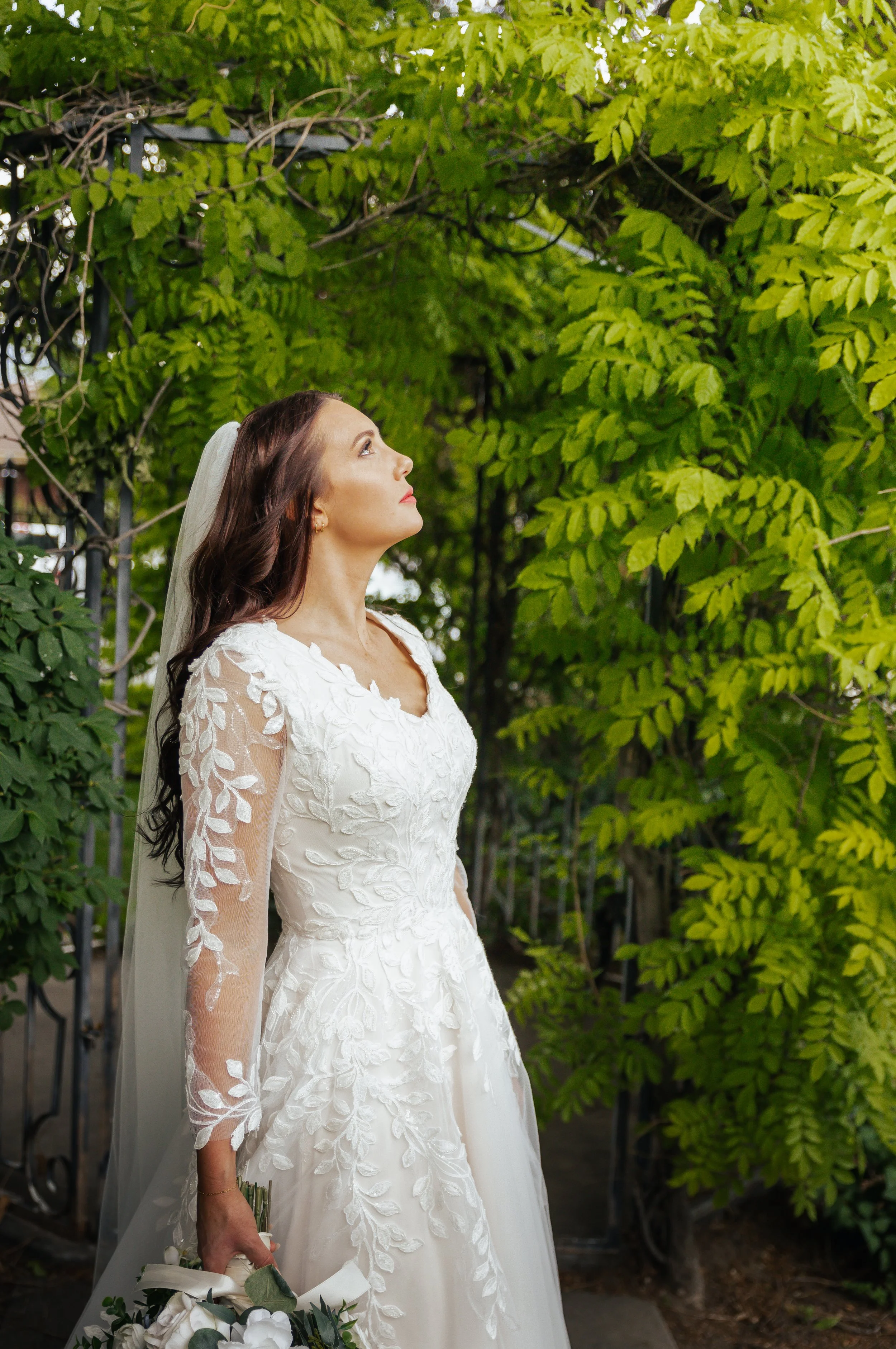 A bride in a white wedding dress with lace details stands outdoors among green foliage, holding a bouquet of white flowers with greenery, looking upward.