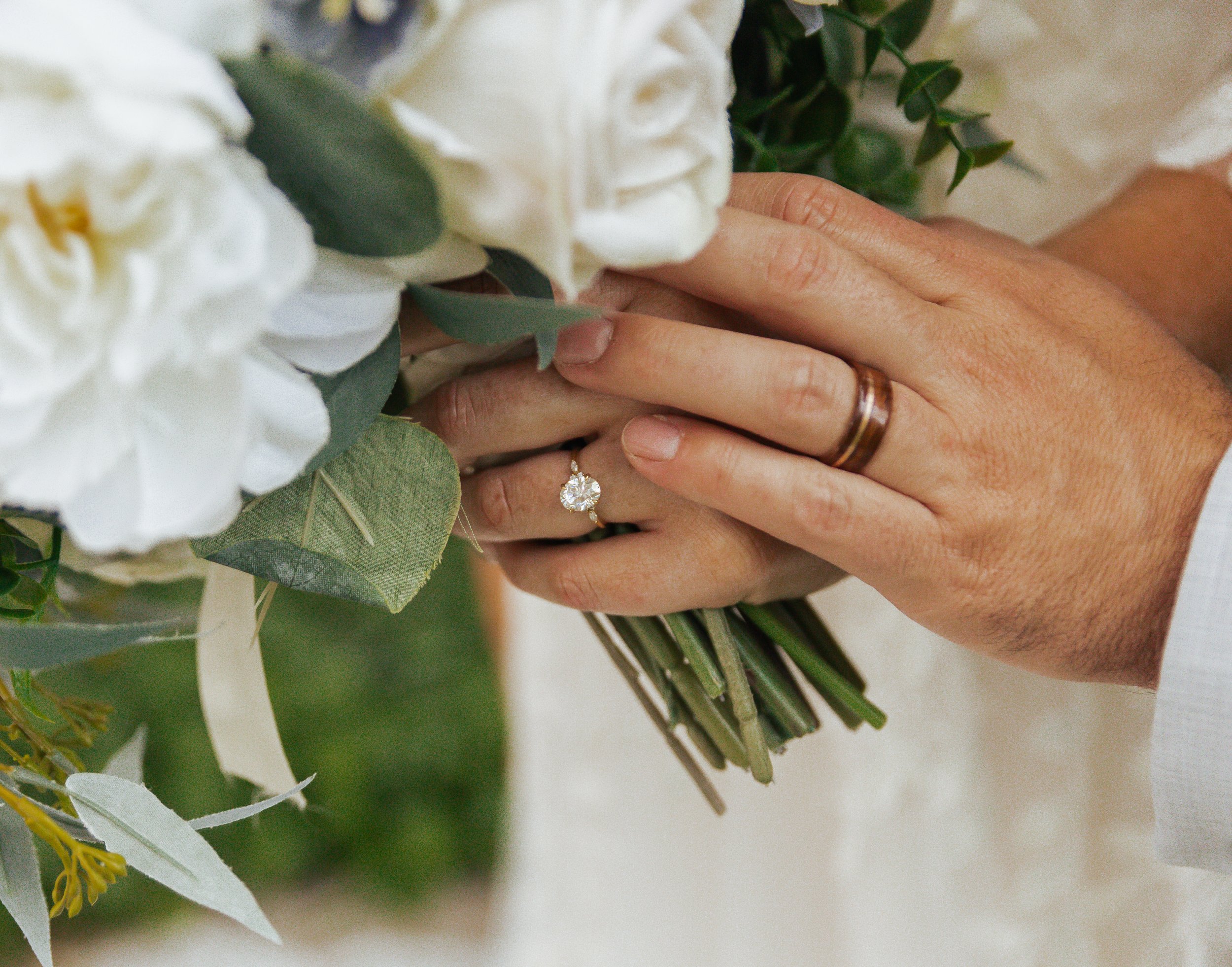 Close-up of hands holding a bouquet of white flowers and green leaves, with a diamond engagement ring on the woman's finger and a wedding band on the man's finger.