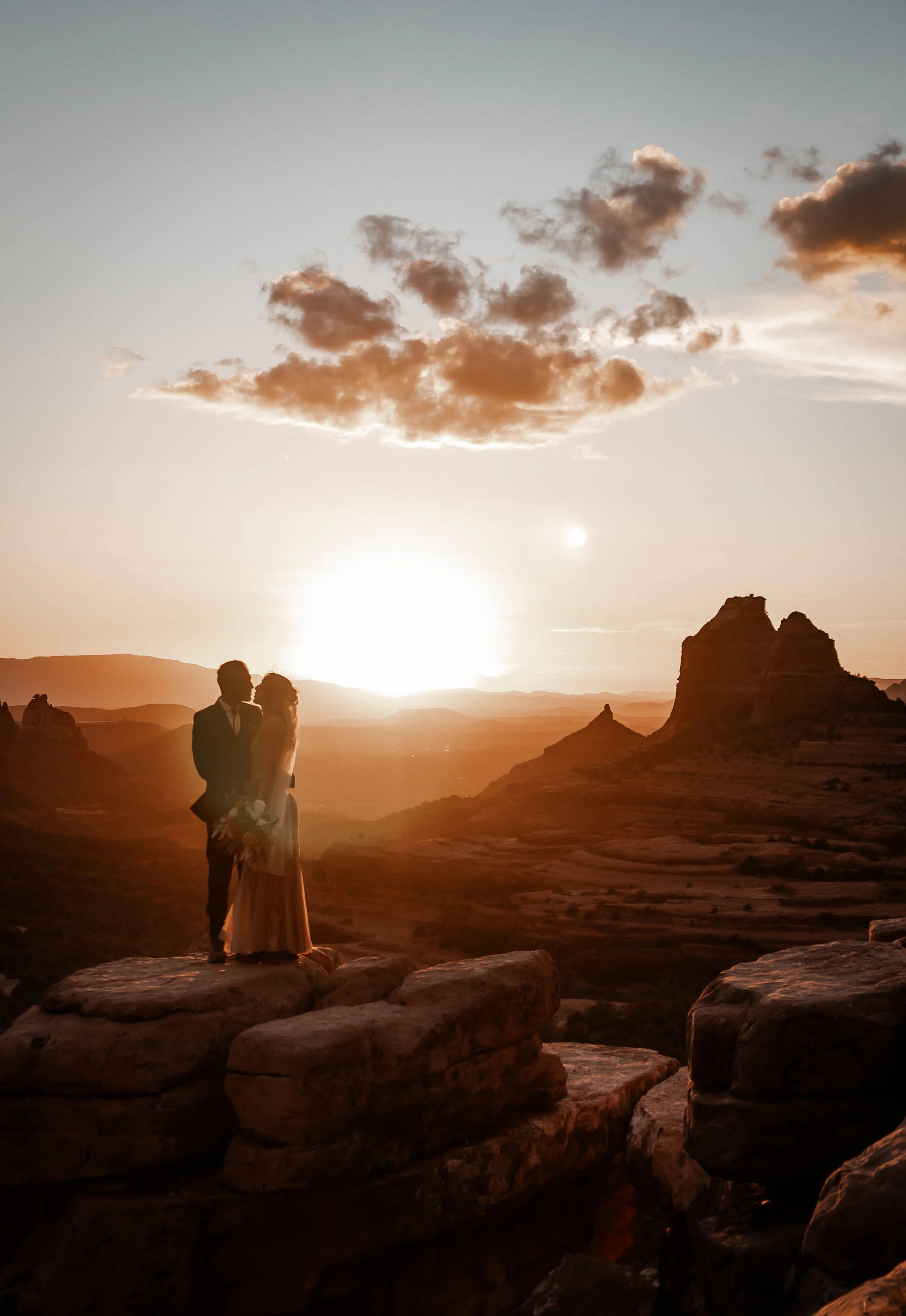 A couple in wedding attire standing on rocks during sunset, with a scenic landscape of mountains and clouds in the background.