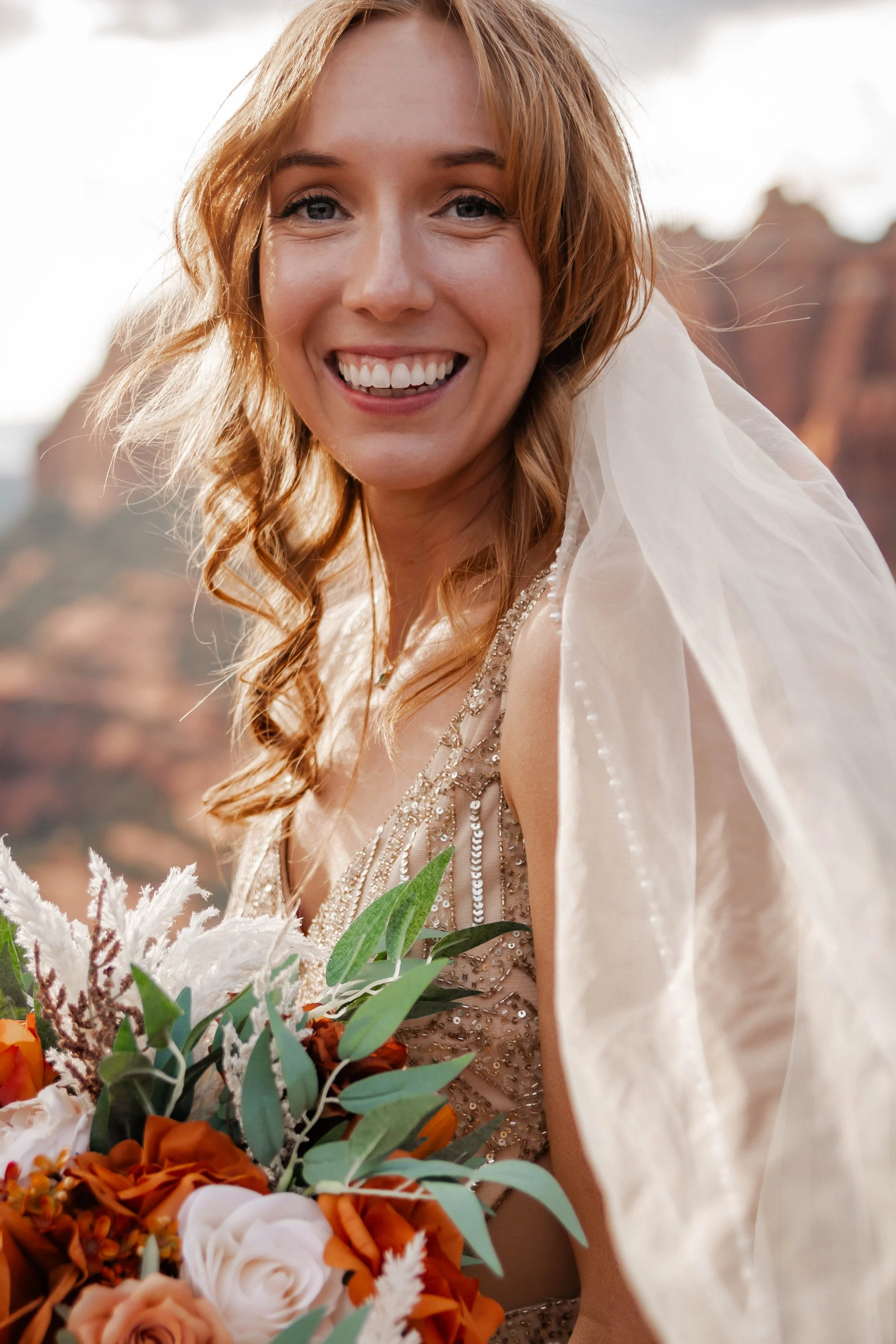 A smiling red-haired bride in a beaded wedding gown holding a bouquet of orange, white, and green flowers, outdoors with a scenic desert landscape in the background.