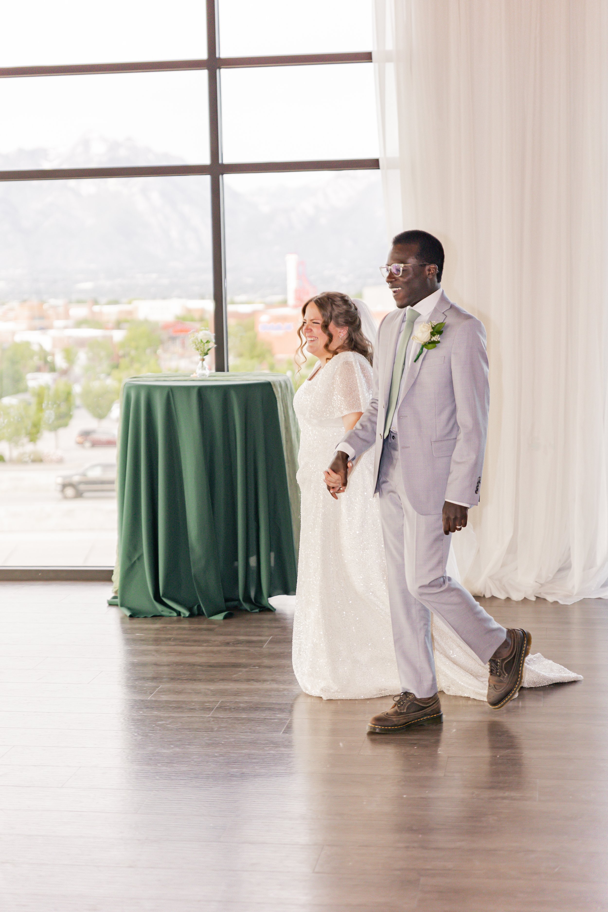 A couple in wedding attire holding hands at their wedding ceremony, smiling and walking across a wooden floor with large windows and a cityscape view in the background.