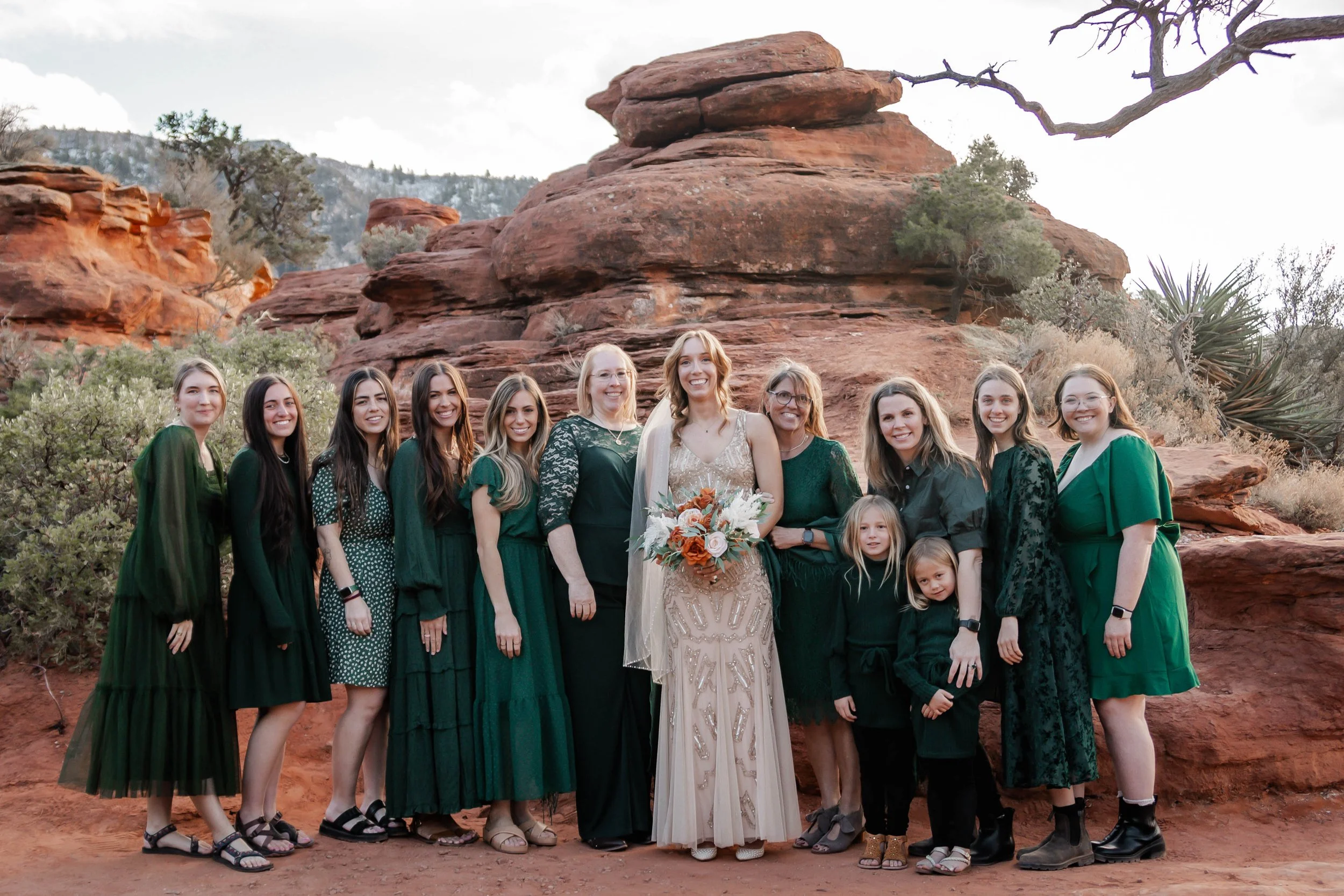 A bride in a beige wedding dress holding a bouquet of flowers, surrounded by women and children in green and black dresses, standing outdoors on red rocky terrain with trees and a large rock formation in the background.