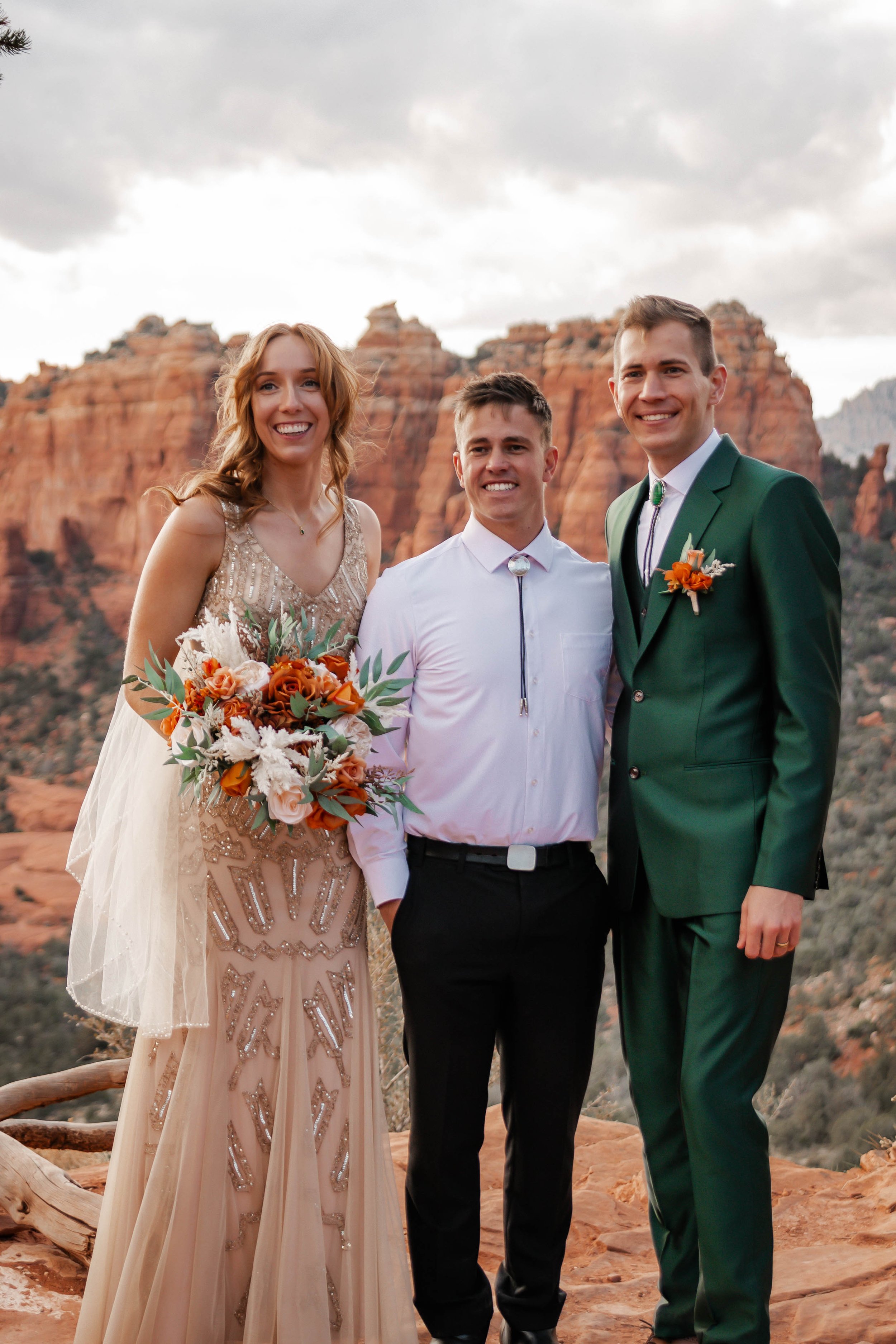 A bride and groom with a man in the middle standing on red rock terrain with red rock formations in the background, dressed in wedding attire, during sunset or late afternoon.