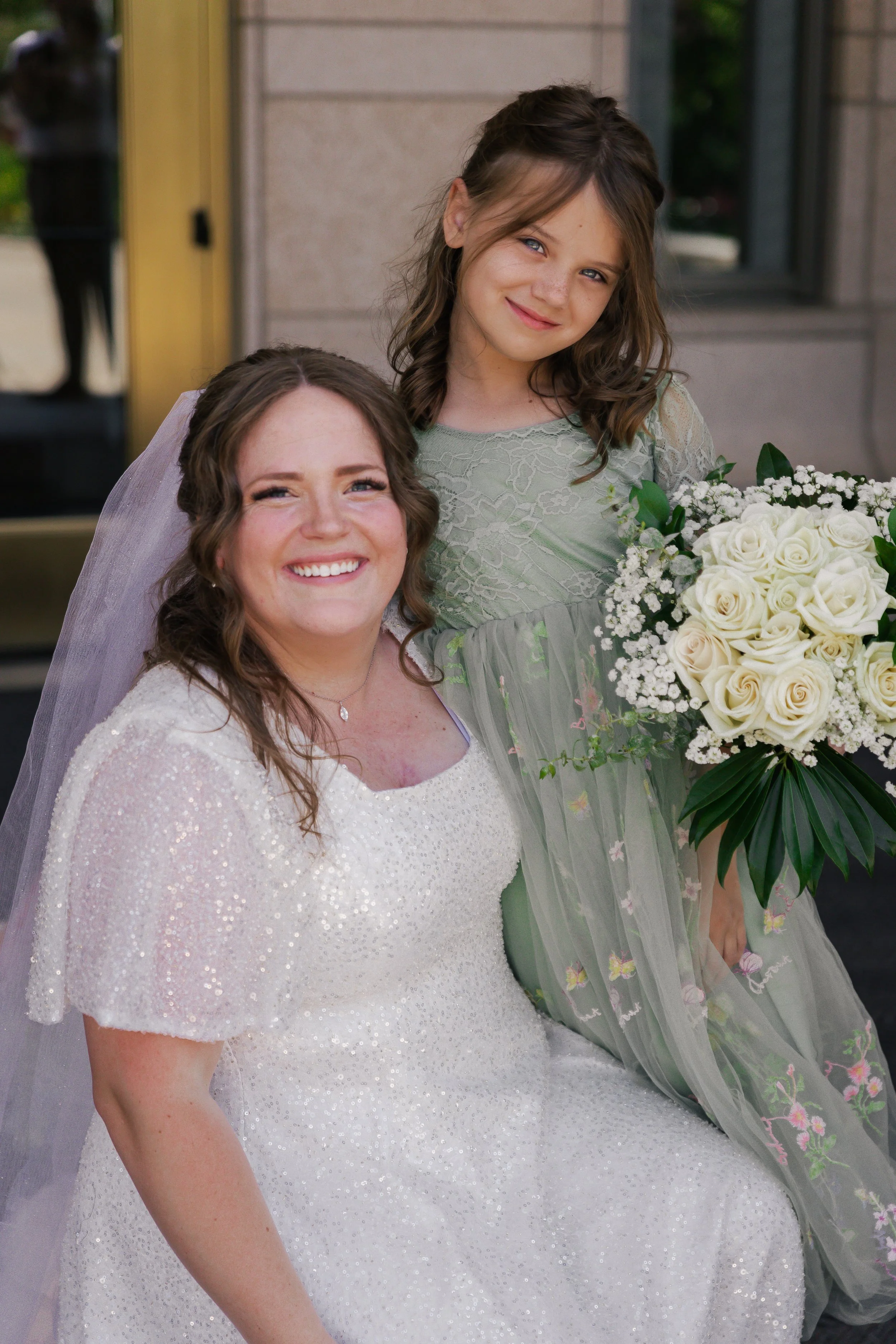 A woman in a wedding dress, sitting and smiling, with a young girl in a green dress holding a bouquet of white roses, both posing outdoors.