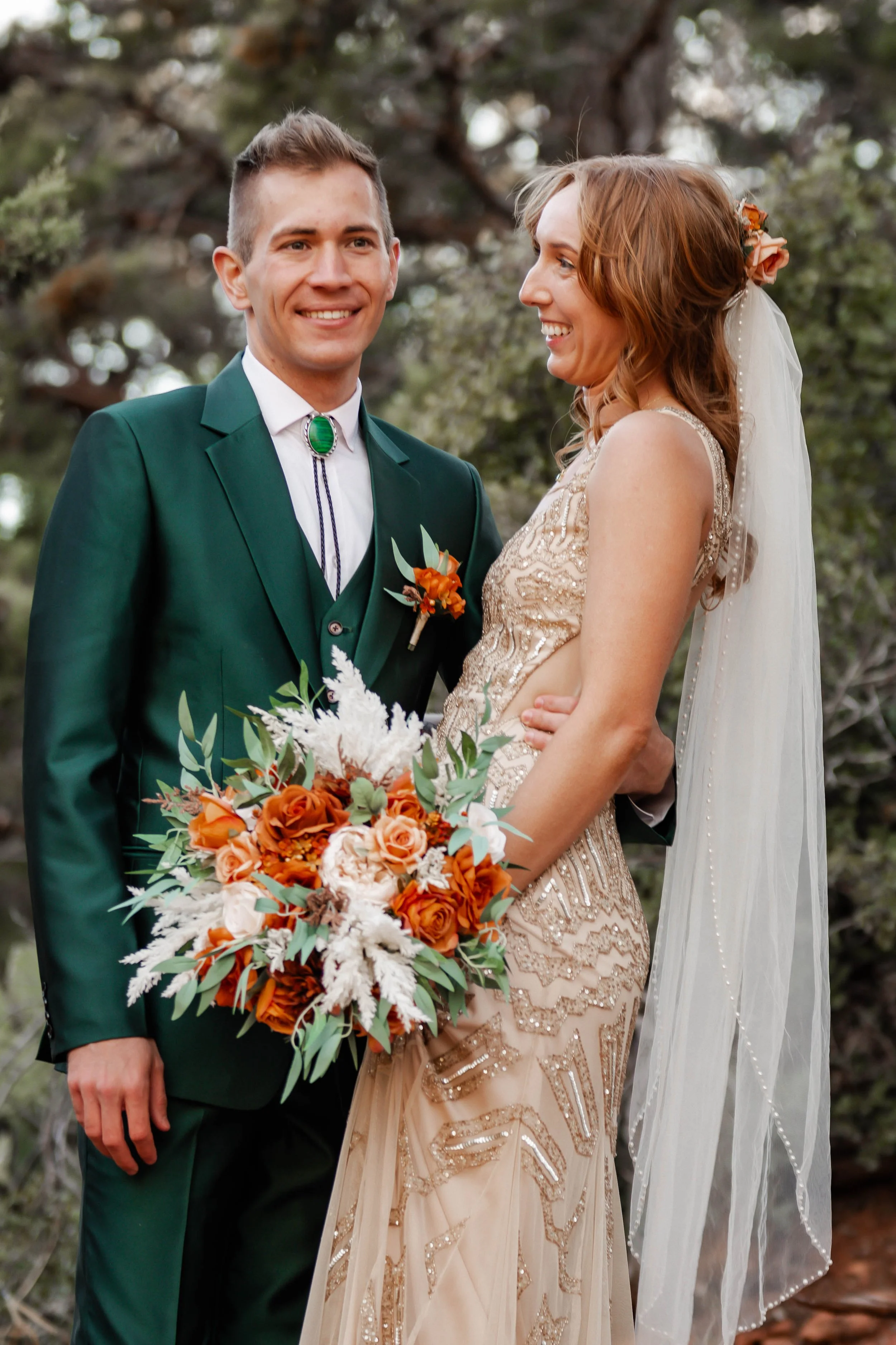 A happy newlywed couple on their wedding day outdoors, with the groom in a dark green suit and the bride in a cream-colored dress holding a bouquet of orange and white flowers.