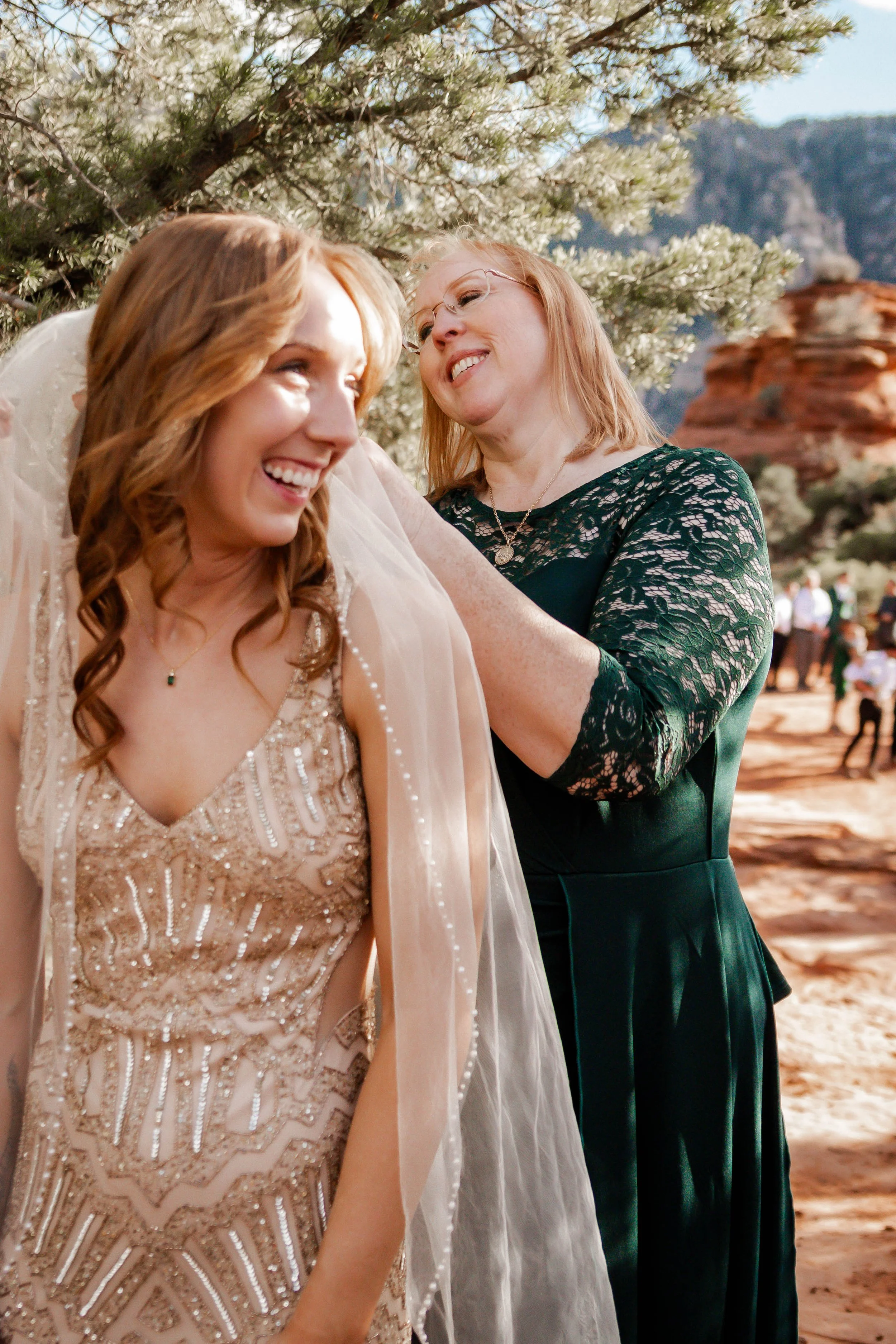 A woman wearing a beaded bridal gown and veil is smiling as another woman adjusts her veil outdoors in a desert landscape with rocks, mountains, and a tree.