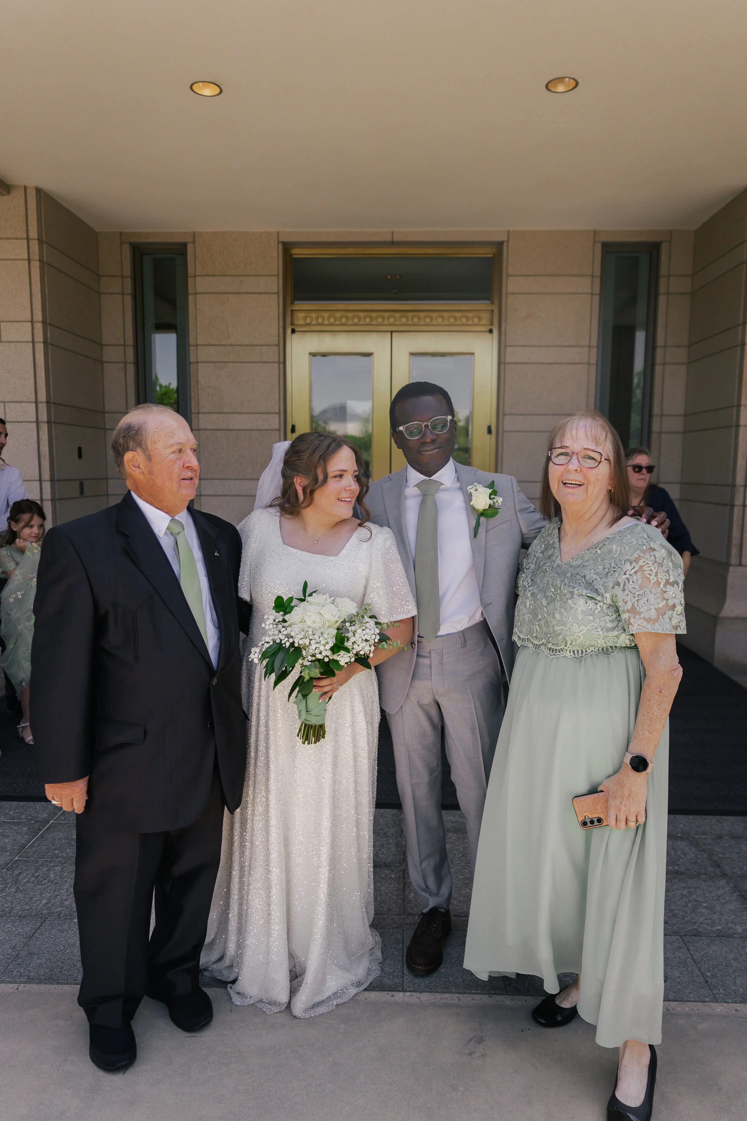 A group of five people standing on the front steps of a building during a wedding celebration. The bride holds a bouquet of white flowers, wearing a white dress, alongside the groom in a light gray suit. Two older women and a man are standing with th