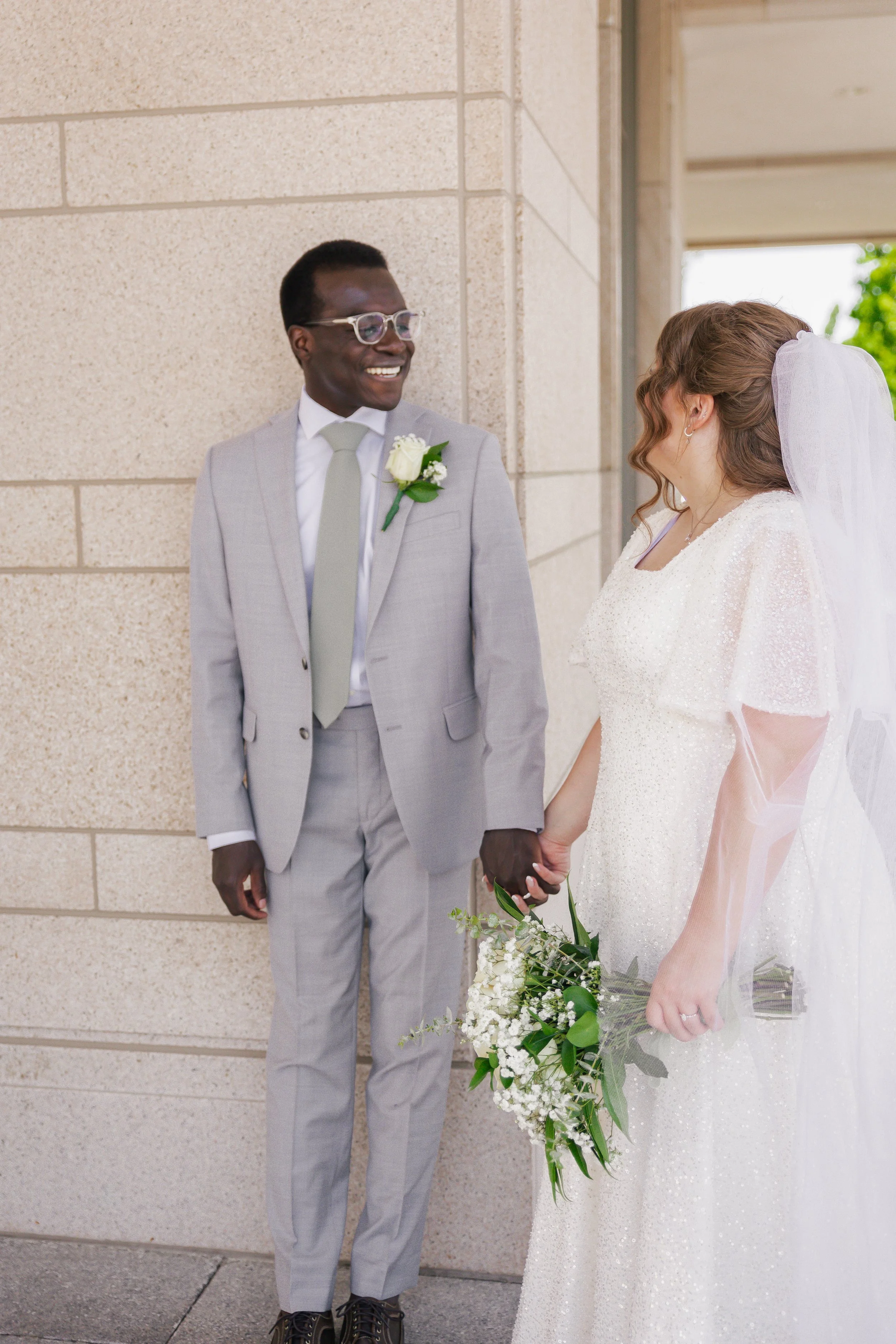 A smiling interracial couple dressed in wedding attire, holding hands in front of a beige stone wall, with the woman holding a bouquet of white flowers.