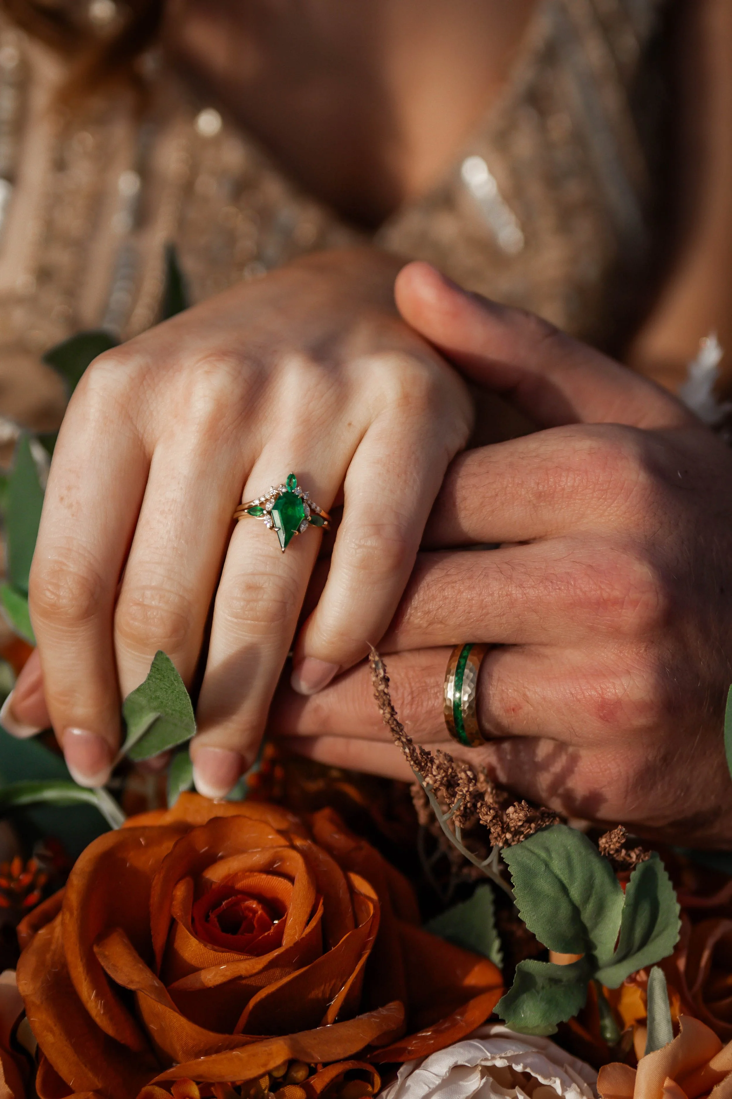 Close-up of a couple's hands showing their wedding rings, with the bride wearing an emerald and diamond ring, and the groom wearing a green and silver band, surrounded by flowers and greenery.