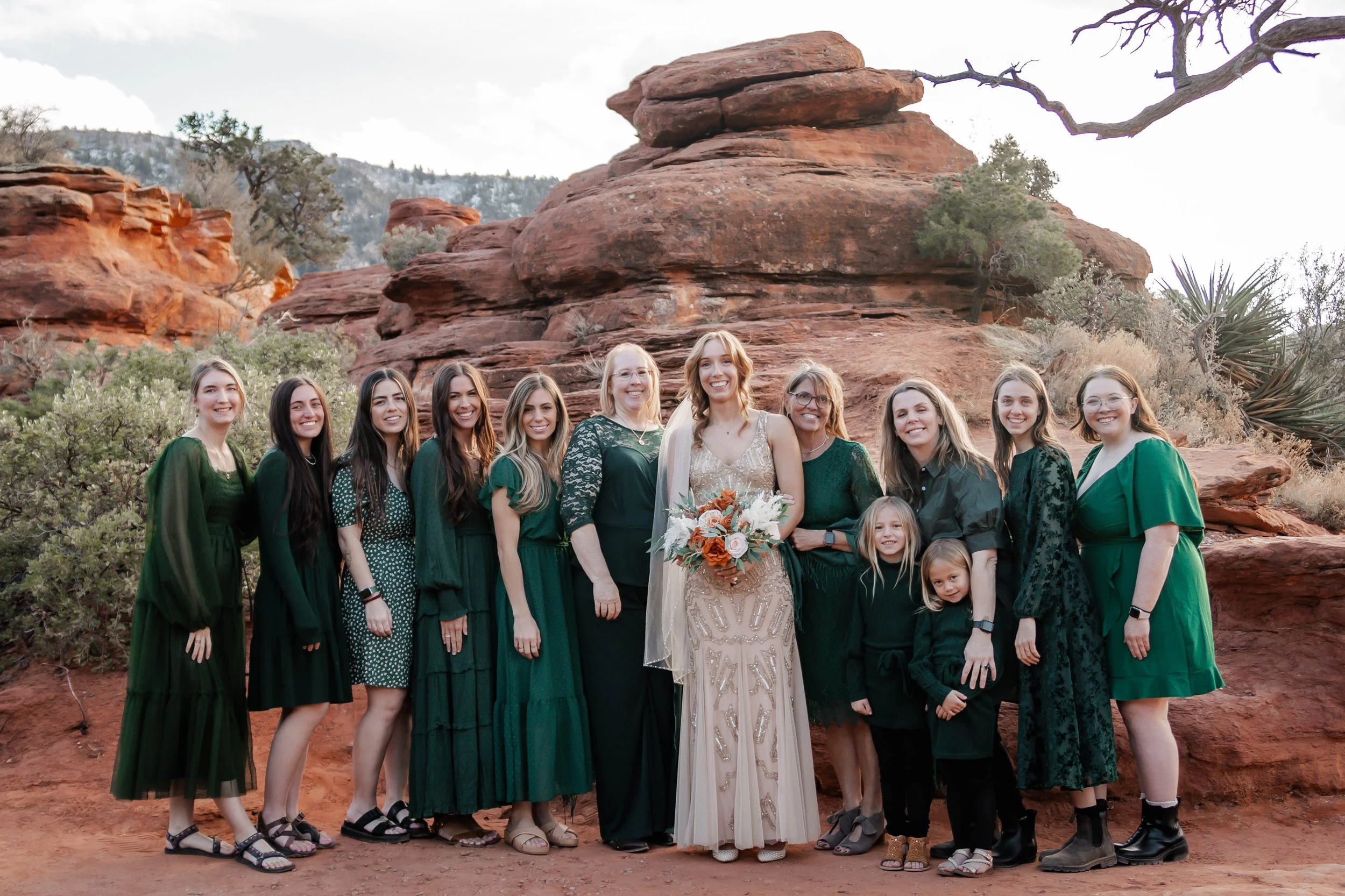 A group of 13 women and 2 young girls standing outdoors in front of red rock formations and desert vegetation, dressed in green and black, celebrating a wedding.