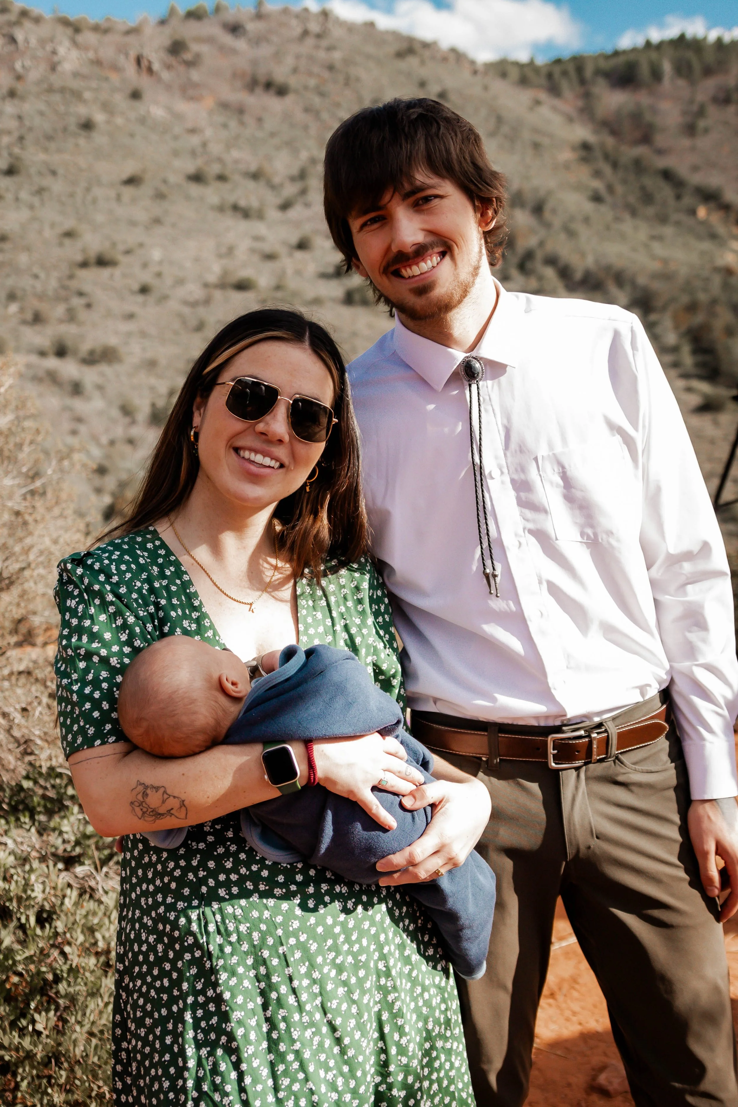 A woman in sunglasses holding a baby, standing next to a man in a white shirt outdoors in a dry, hilly landscape.
