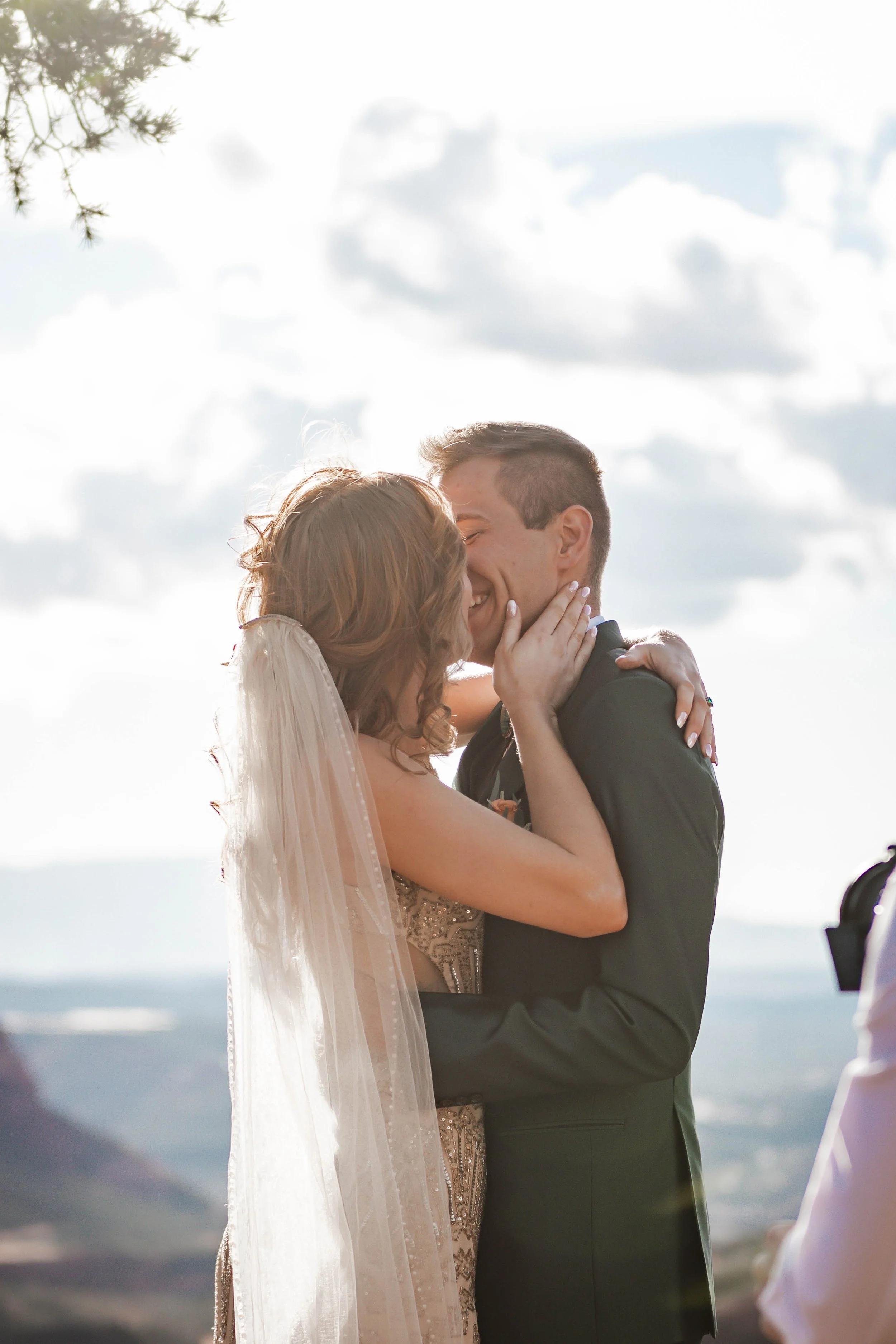 A bride and groom sharing a kiss during their wedding outdoors against a backdrop of clouds and mountains.