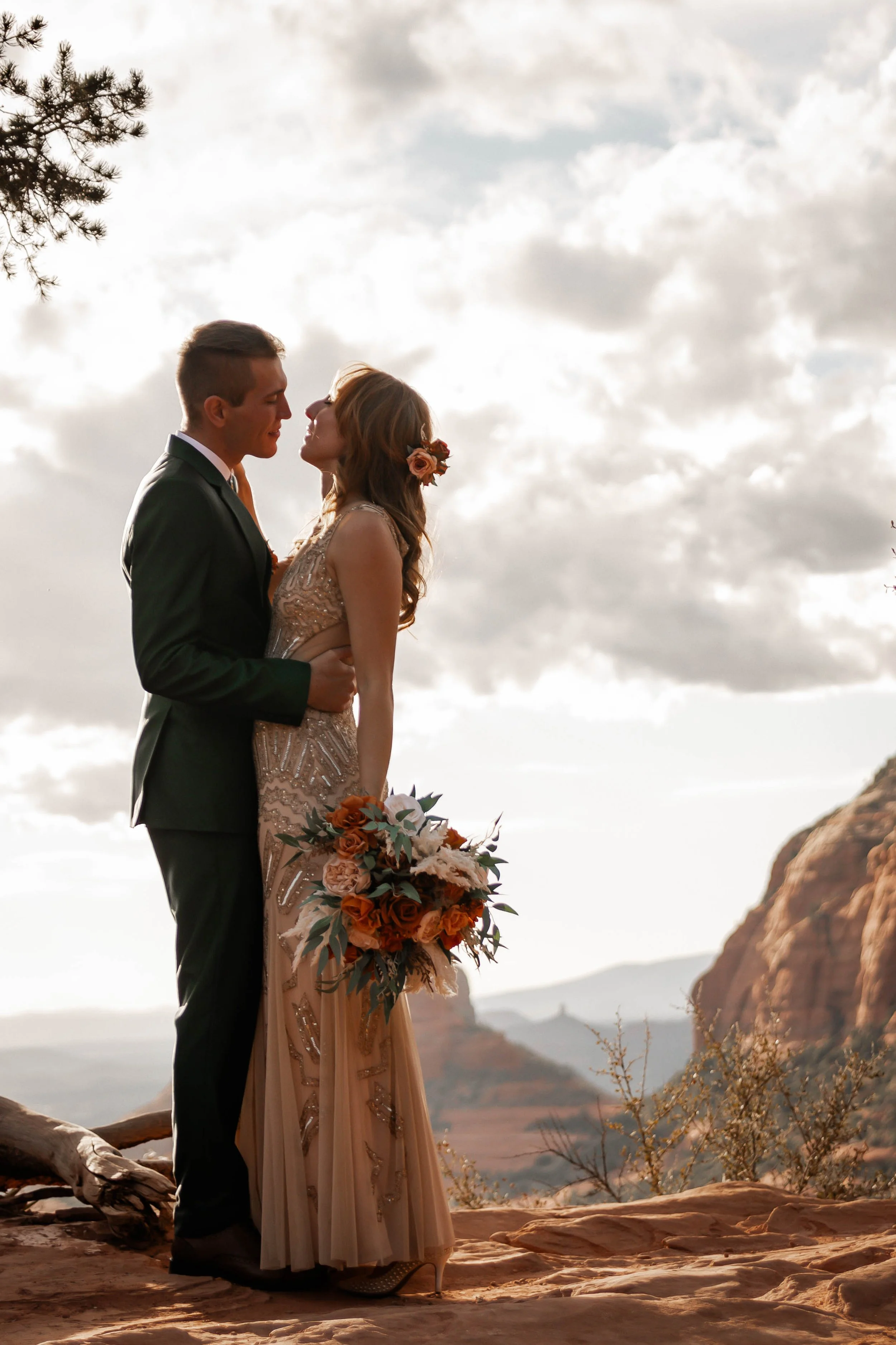 A couple in formal attire about to kiss outdoors on a rocky landscape with mountains and cloudy sky in the background, the woman holding a large bouquet of orange, white, and green flowers.
