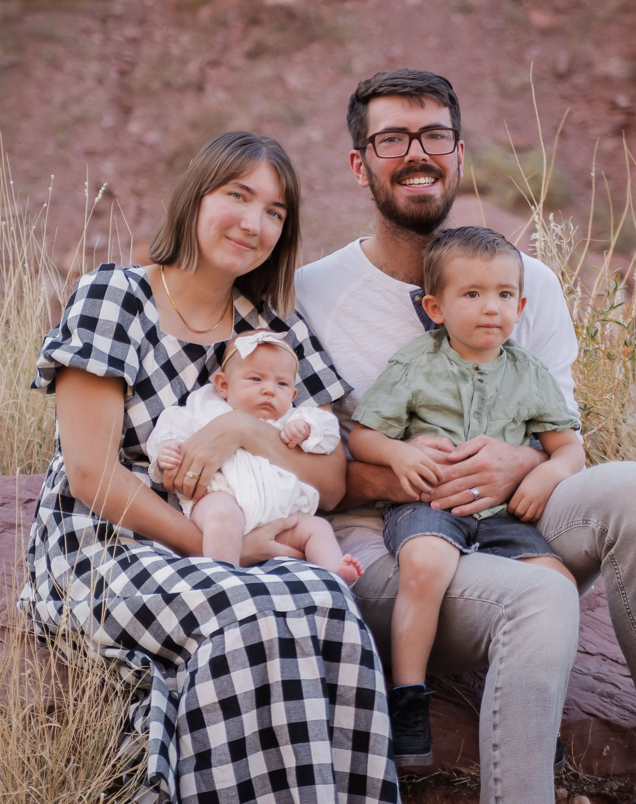 A family of four sitting outdoors on a rocky landscape with dry grass and pinkish rock background. The mother holds a baby girl with a bow in her hair, the father sits next to a young boy. The family is smiling.