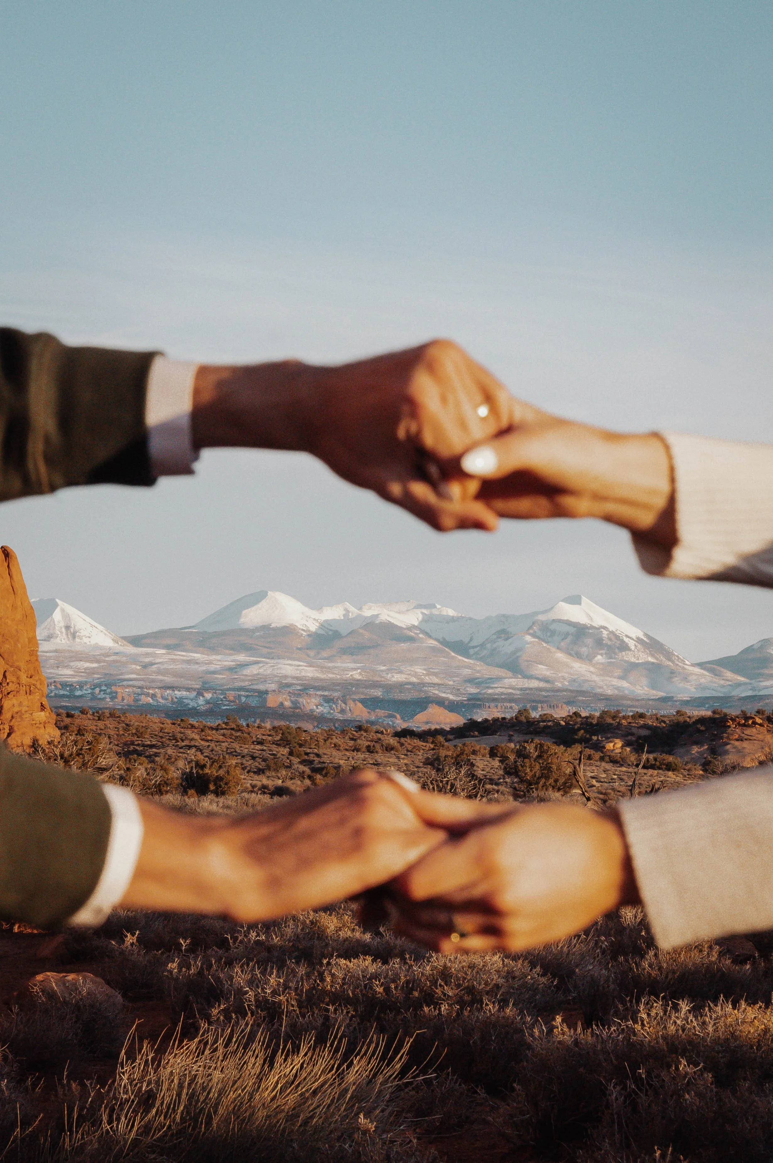 Two pairs of hands holding each other in a desert landscape with snow-capped mountains in the background.