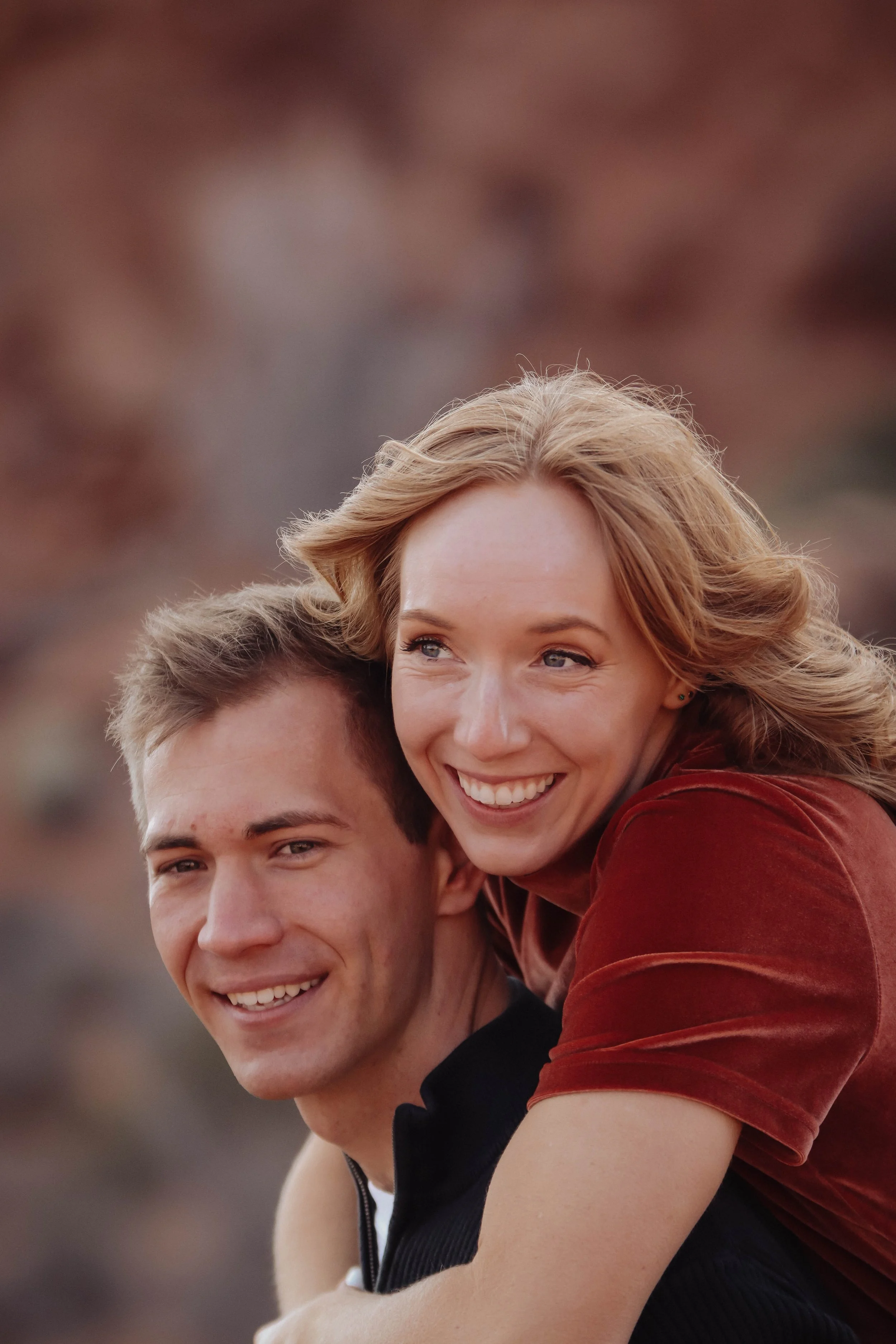 A woman giving a piggyback ride to a smiling man outdoors with a blurred earthy background.