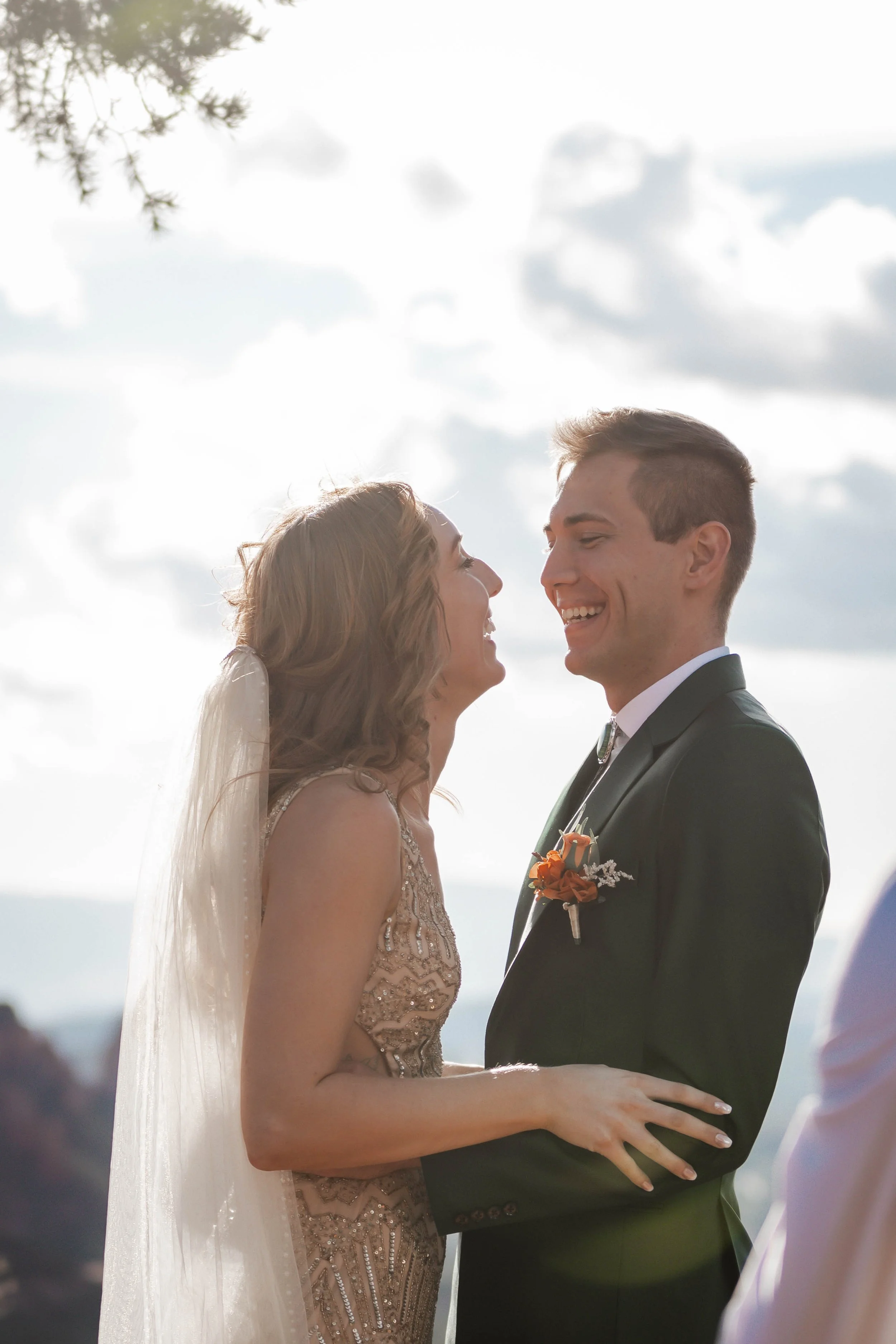 A bride and groom smiling and looking at each other during their wedding ceremony outdoors with a cloudy sky and trees in the background.