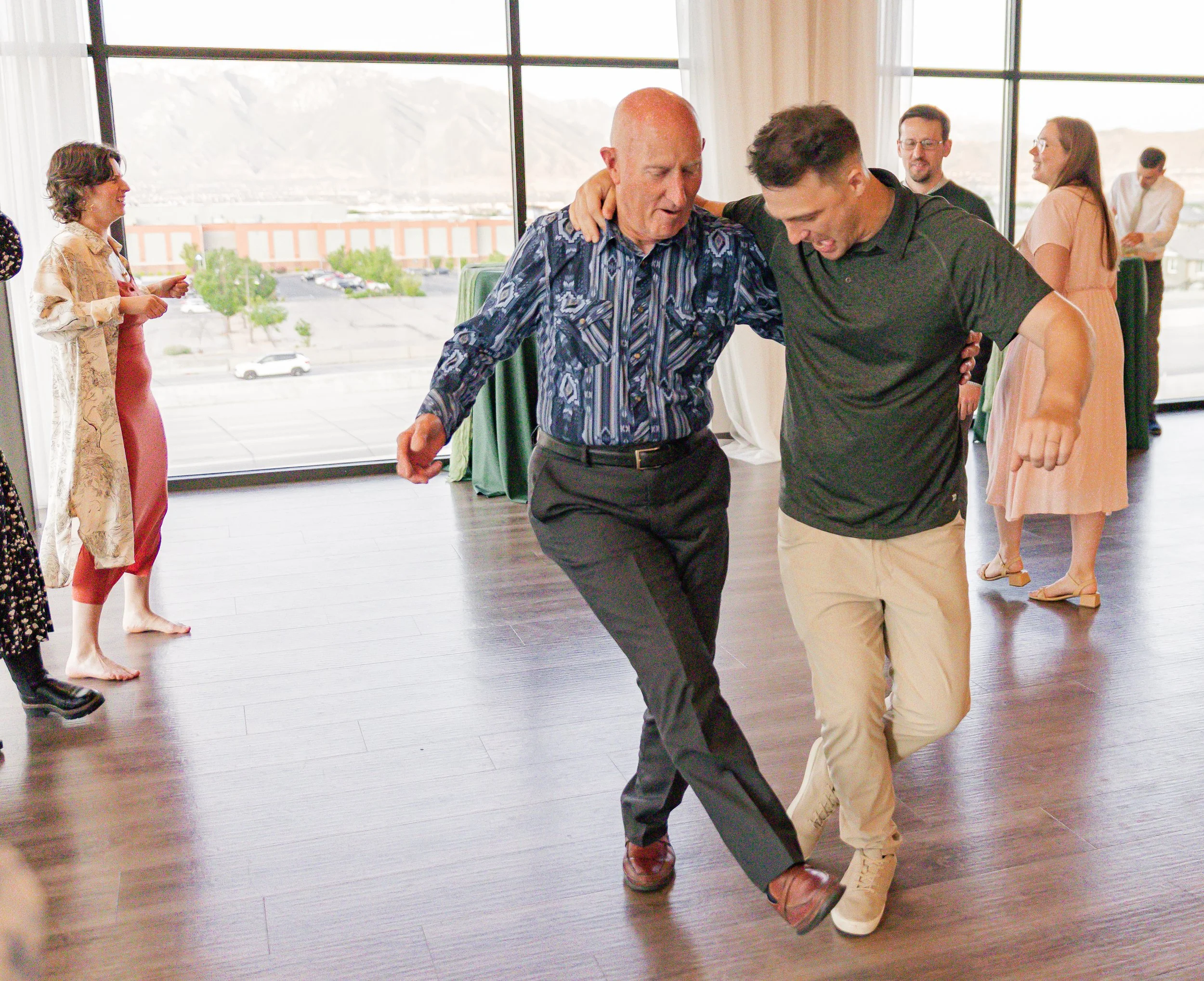 Two men dancing together on a wooden floor in a room with large windows, with other people standing and chatting in the background.